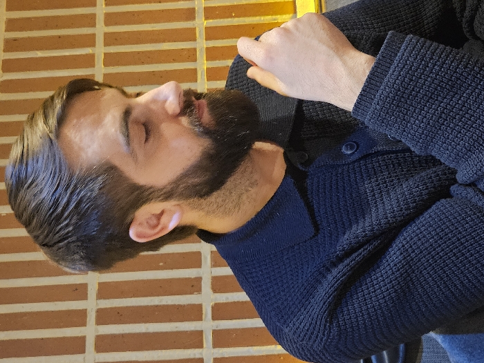 A man with dark hair and a full beard is seen from the side. His eyes are closed. He wears a navy blue collared shirt with a cardigan on top. The cardigan is knitted and has buttons. His left hand rests on his chest. The background features orange and brown tiles arranged horizontally. A thin, yellow line appears at the top of the image, likely from a light source.