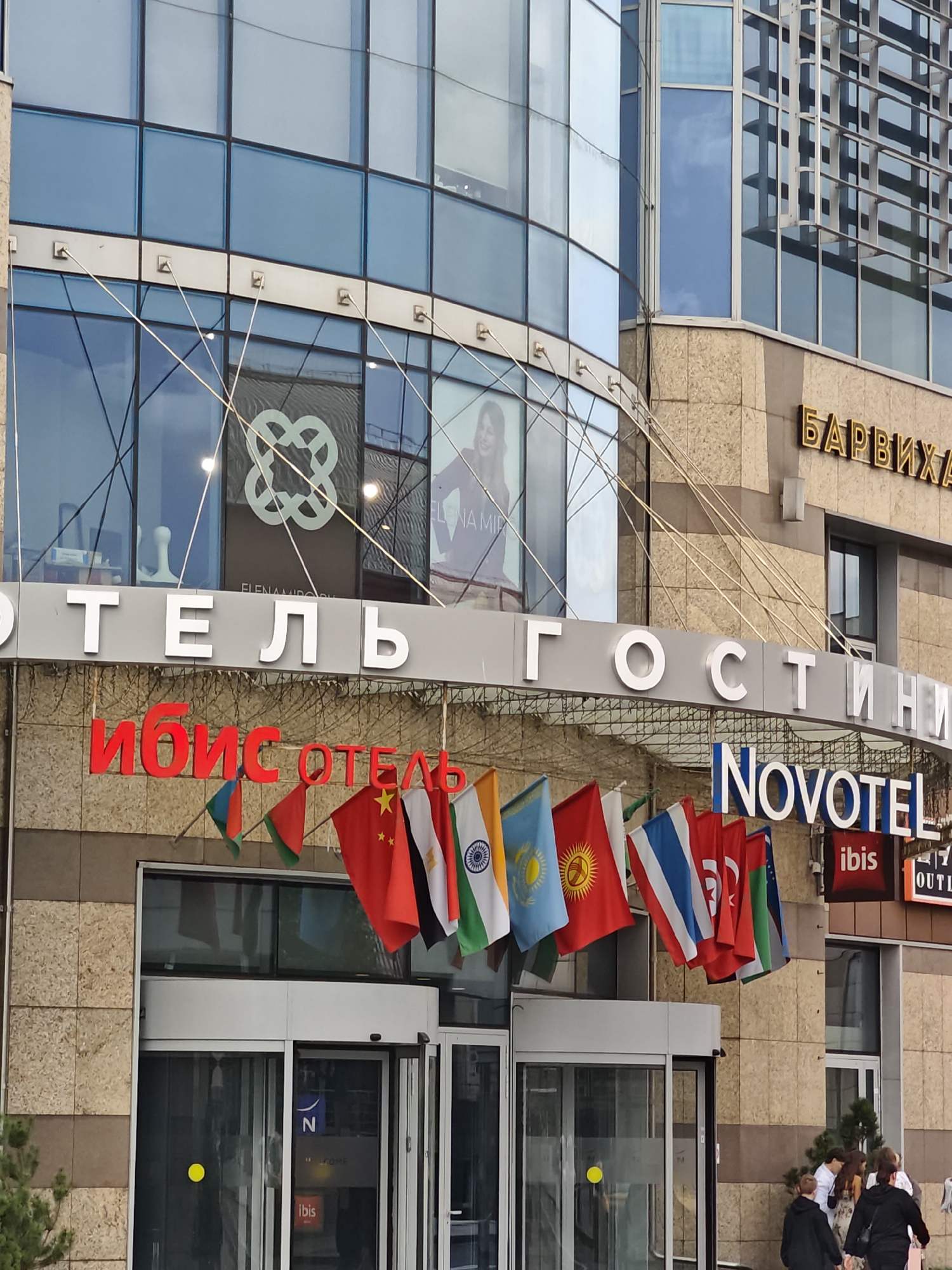 The image shows the exterior of a multi-story building in Moscow, Russia. The building has a modern facade with large glass windows and stone cladding. Prominently displayed are signs for "ОТЕЛЬ" (Hotel) and "ГОСТИНИЦА" (Guesthouse) in large white letters. Below these, a red sign reads "ИБИС ОТЕЛЬ" (Ibis Hotel). The flags of various countries are displayed on a string above the entrance. A Novotel logo is visible to the right of the "ГОСТИНИЦА" sign. Several people are walking in front of the building. The overall scene suggests a commercial or hospitality establishment.