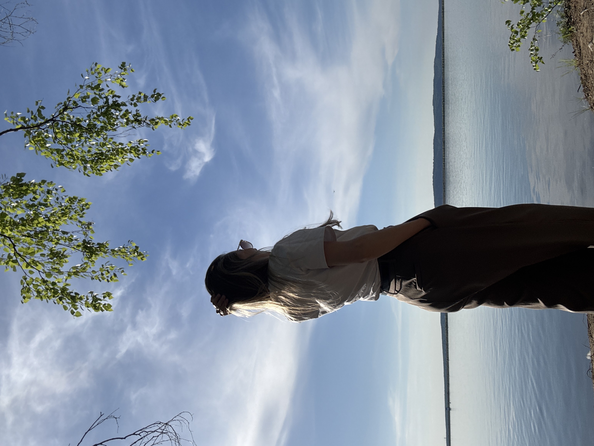 The image shows a woman standing near a body of water, likely in a natural setting. She is wearing a white t-shirt and dark pants. Her back is facing the water, and she is looking up at the sky. The sky is blue with some wispy clouds. There are trees visible on either side of the frame. The image was taken in the city of Chaikovsky, Russia.