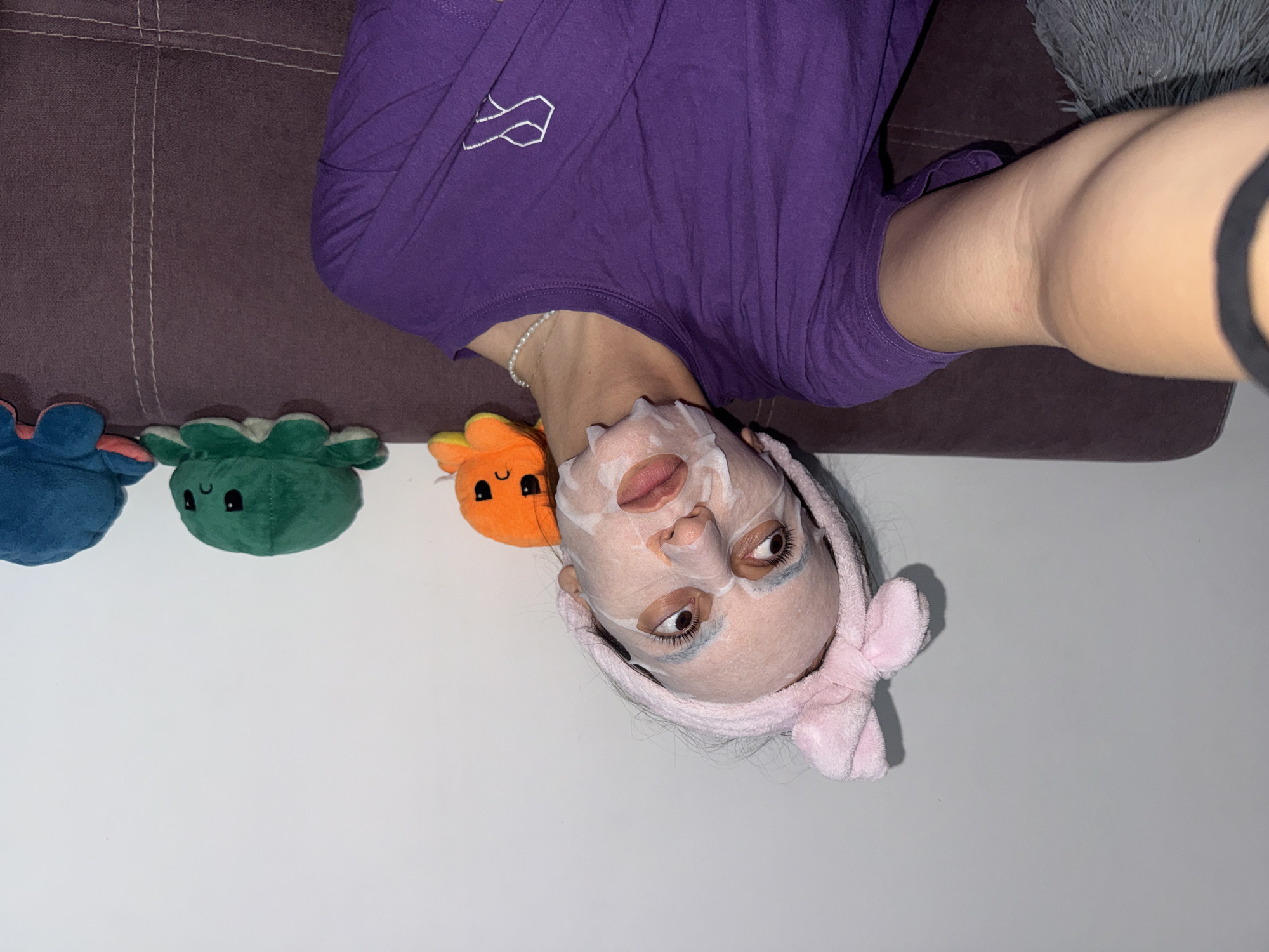 A young woman lies on a brown couch, wearing a purple t-shirt, a pearl necklace, and a pink towel headband with a bow. She has a sheet mask on her face. Next to her on a white surface are three plush toys: a blue octopus, a green octopus, and an orange plush. The location is not identifiable as Plovdiv, Bulgaria.
