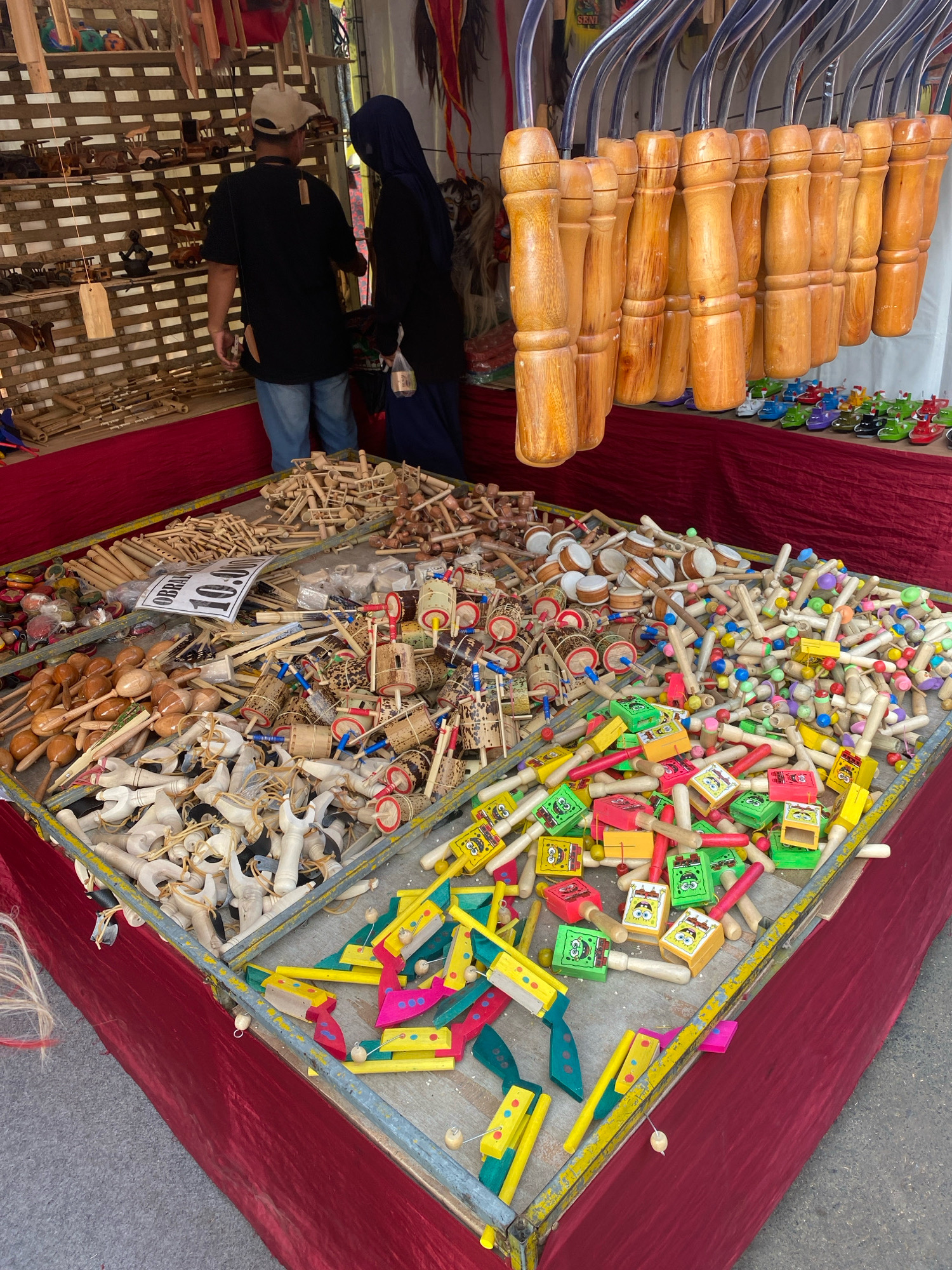 At a street market stall in Nyamok, Indonesia, a variety of handcrafted wooden toys are displayed. The stall features a central table overflowing with small wooden objects. These include numerous miniature drums with colorful accents, wooden maracas, and carved wooden figures, some resembling horses and others more abstract. Scattered amongst these are toy spinning tops, small wooden boxes adorned with cartoon characters, and simple wooden puzzles. In the background, shelves are stocked with more wooden items, including what appear to be model vehicles and other decorative pieces. Two individuals are visible in the upper left portion of the image, examining the merchandise. A man wearing a baseball cap and a dark t-shirt stands facing the shelves, while a woman in a dark hijab and black garment is beside him, looking at the displayed items. The stall is draped with a deep red fabric, suggesting a festive or artisanal atmosphere typical of local markets.
