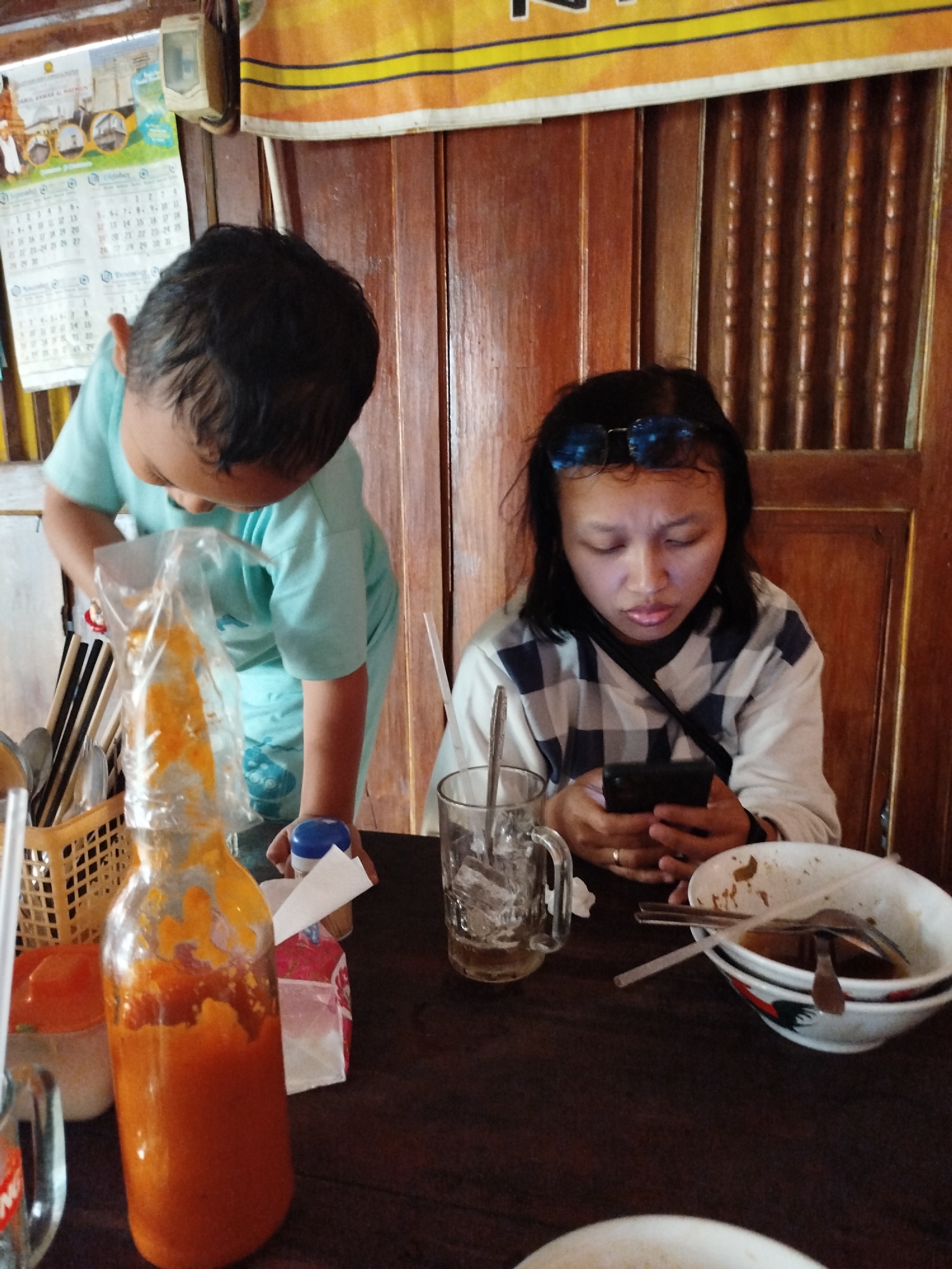 In a dimly lit room with wooden paneled walls, a young child in a light blue t-shirt leans over a table, seemingly curious about a bottle of orange sauce. The bottle, partially covered in plastic, sits on the dark wooden surface. To the right, a woman in a plaid shirt stares intently at her smartphone, her glasses perched on her forehead. The table is cluttered with various items, including a basket of chopsticks, an empty glass, and a bowl with remnants of food. A calendar hangs on the wall in the background, hinting at a domestic or casual setting, potentially in Pojok, Indonesia.