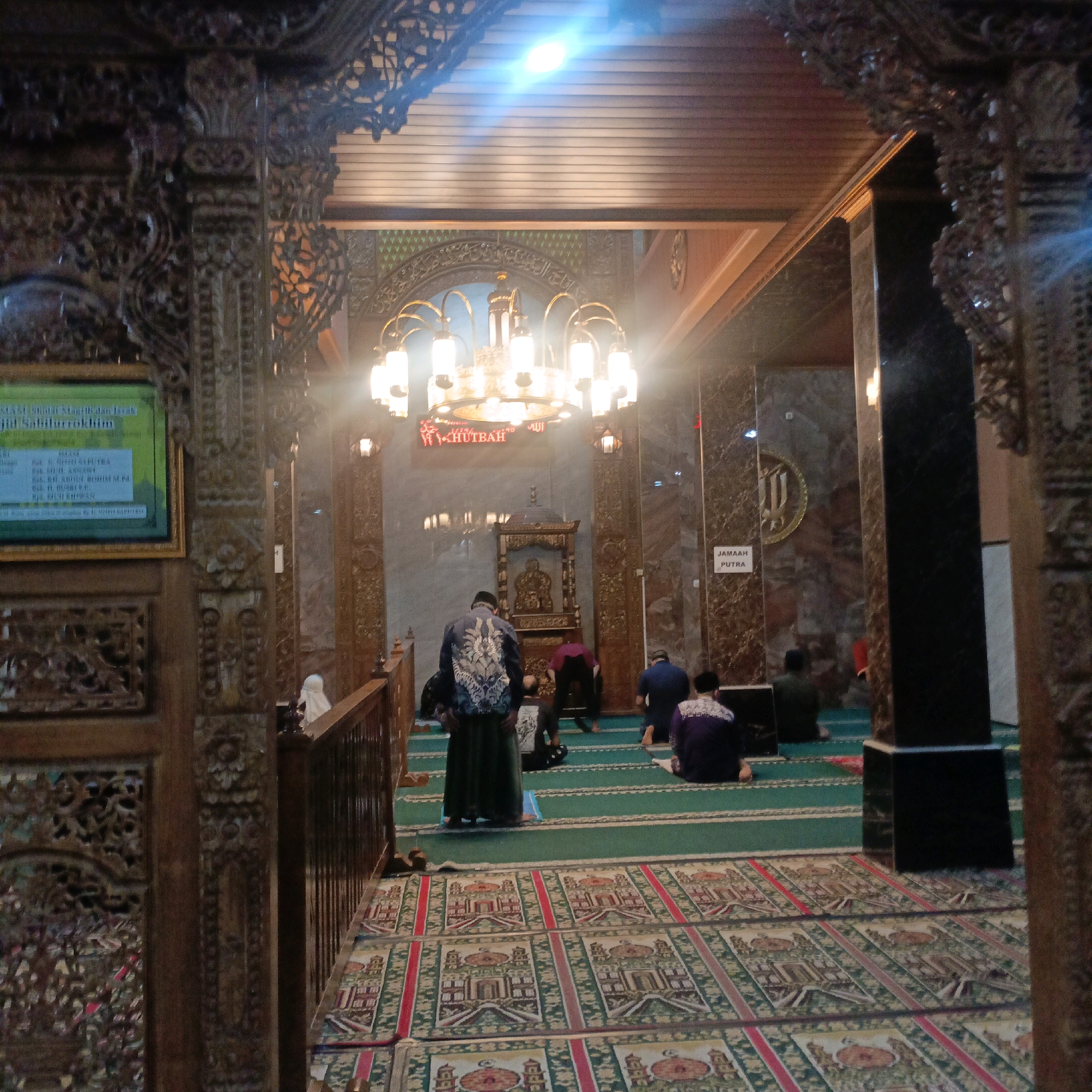 This image depicts the interior of the Masjid Sabilurrokhim, located in Salatiga, Indonesia. The mosque features ornate wooden carvings on the pillars and ceiling, and a large chandelier hangs prominently. Several people are seen praying on green prayer mats, which are laid out on a carpeted floor with intricate patterns of mosques and architectural elements. In the background, a raised platform with decorative stonework and a mihrab (a niche indicating the direction of prayer) is visible. A sign indicating "JAMAAH PUTRA" (male congregation) is also present. The lighting suggests it is either early morning or late evening. The scene captures a moment of communal worship within the mosque.