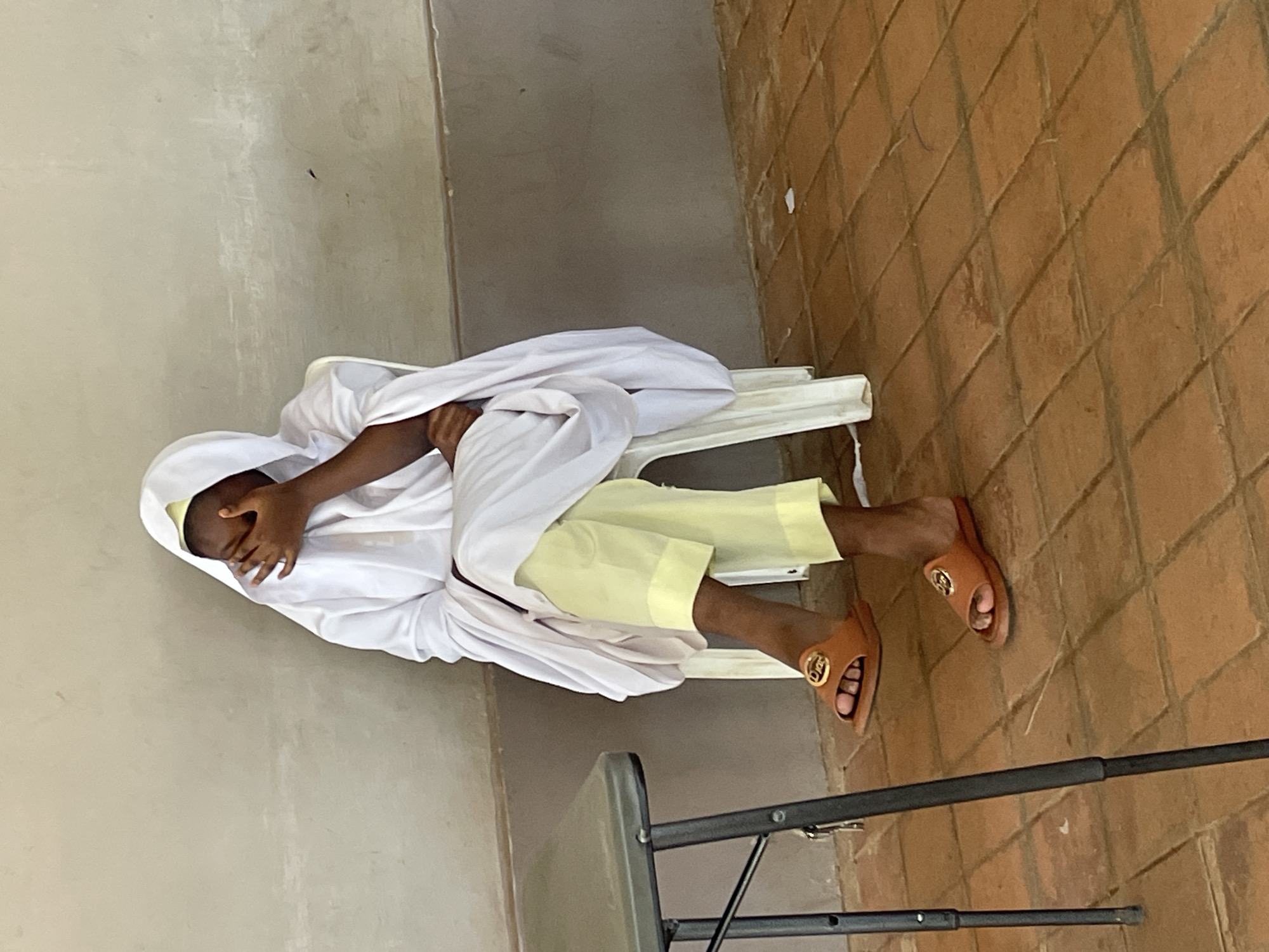 A person wearing a white hooded robe and light yellow pants sits on a white plastic chair in Barakin Lerawi, Nigeria. The person's face is obscured by their right hand. The individual is wearing brown slides with gold embellishments. The background consists of a plain, textured wall and a tiled floor. A dark metal table with a grey top is partially visible in the lower left of the frame.
