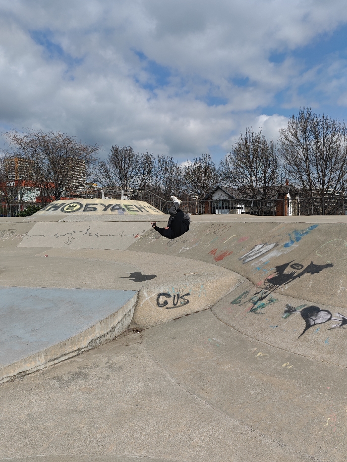 The image depicts a concrete skatepark located in Shumen, Bulgaria, under a partly cloudy sky. A single individual, wearing dark clothing and white athletic shoes, is captured mid-motion, positioned with their torso bent backward and legs raised, appearing to be falling or performing a trick on a curved concrete transition. A distinct shadow of the person is cast on the concrete surface directly beneath them.
The skatepark features various concrete elements, including ramps, ledges, and curved transitions, all marked with graffiti. A prominent curved concrete barrier in the background bears the graffiti "ШУМЕН БУДЕЩЕ" (Shumen Future) in Cyrillic. Other graffiti includes the word "GUS" on a lower ramp and various abstract and stylized drawings. A flat, blue-painted concrete ledge is visible in the lower-left foreground.
In the background, bare deciduous trees line the area behind a metal railing structure. Beyond the trees on the left, multi-story apartment buildings are visible. To the right, smaller, older residential houses with sloped roofs are discernible behind the trees. The scene is illuminated by natural daylight, indicating partial sun with scattered clouds.