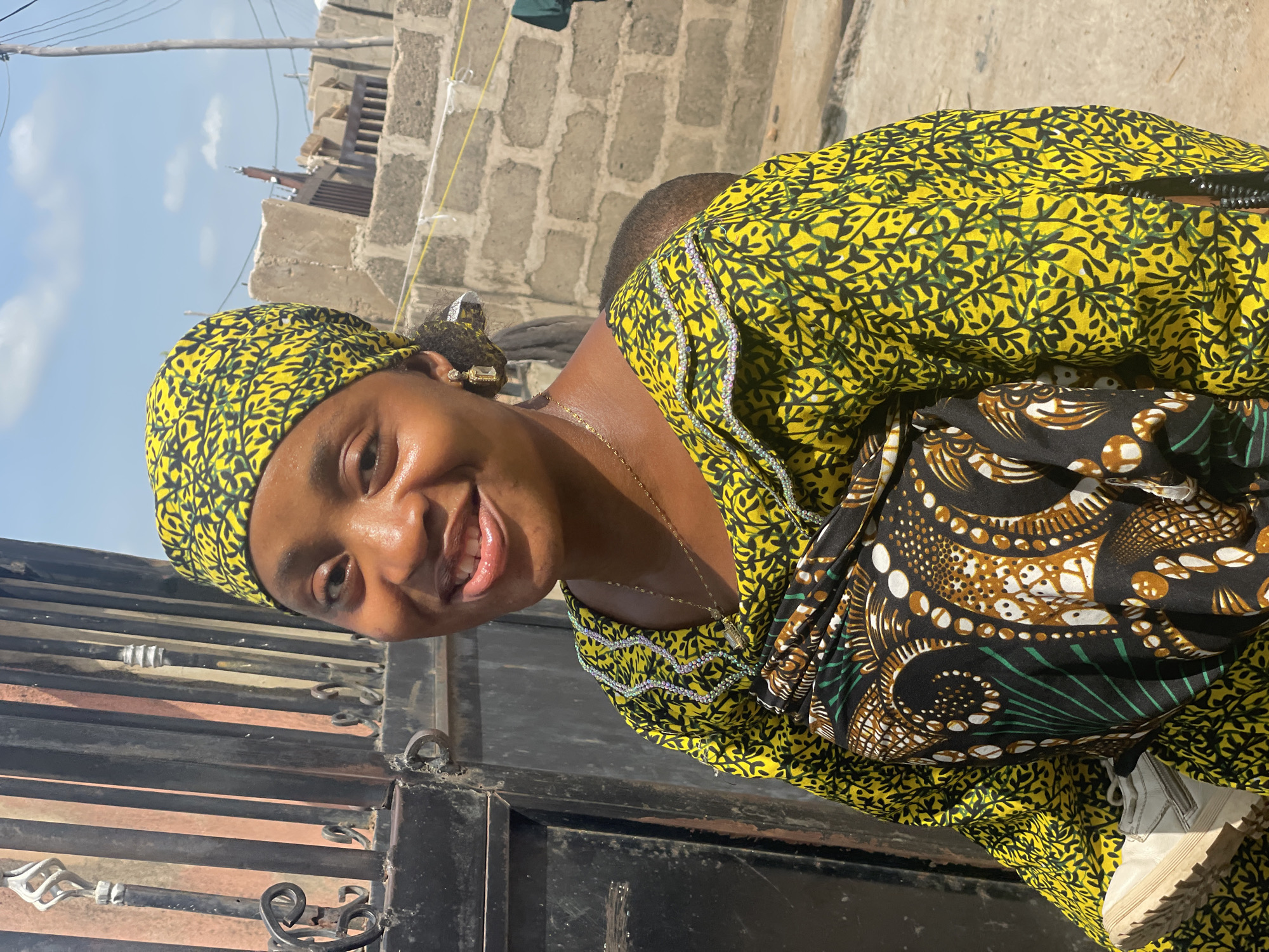 A woman stands outdoors in what appears to be Lokoja, Nigeria, based on the building style and cultural attire. She is fair-skinned, wearing a yellow patterned headwrap and dress with decorative trim around the neckline. A child is bundled on her back using a black and patterned cloth carrier. The woman is smiling and facing the camera. Behind her is a brick structure, power lines, and a partially visible iron gate in the foreground. The sky is blue.