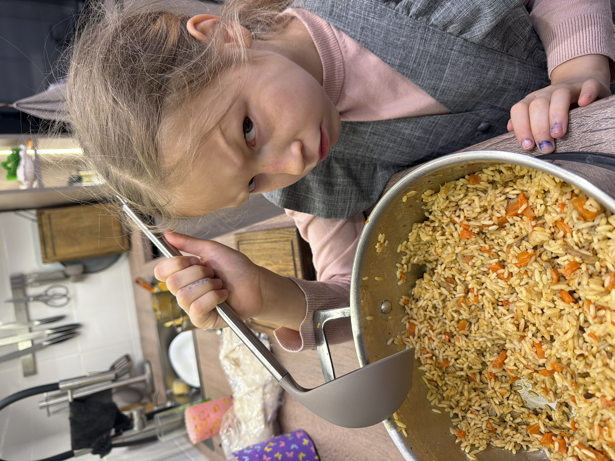 A young girl is closely inspecting a pot of pilaf with a ladle in her hand. She is wearing a pink sweater and a grey vest. Her hair is blonde, tied in a ponytail. The pilaf is cooked with rice and carrots. The background shows a kitchen setting with various utensils and items on the counter. The location is Dubovaya, Russia.