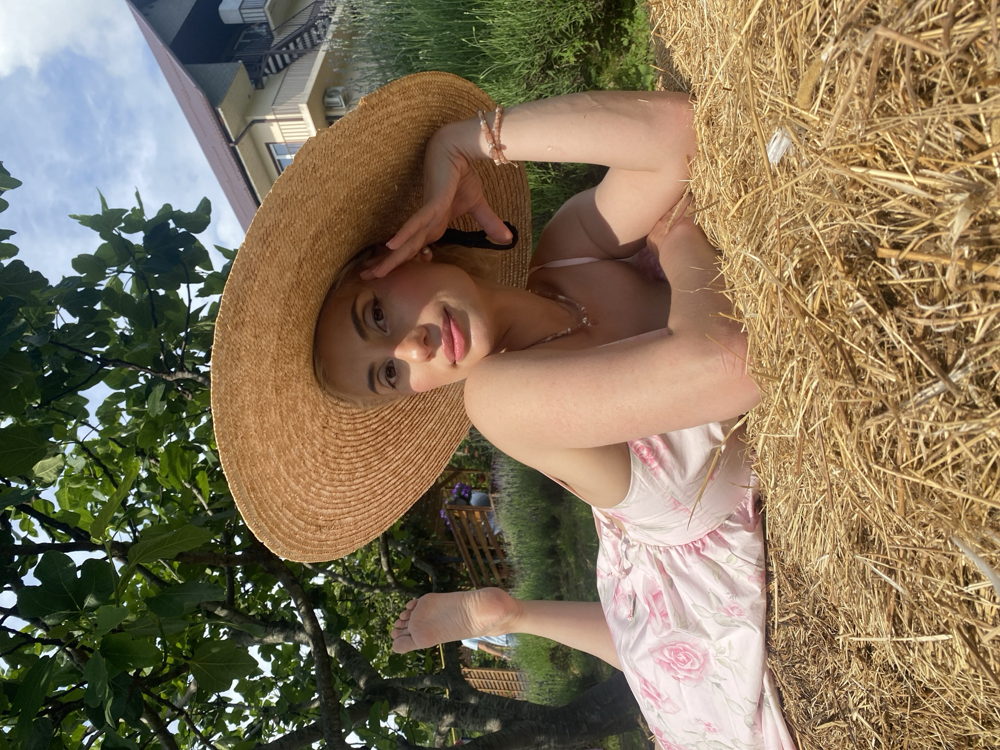A woman in a wide-brimmed straw hat and a pink floral dress lies on a bale of hay in a garden in Verkhneye Uchdere, Russia. The woman is propped up on her elbows, her left leg bent with her foot in the air. She is wearing jewelry, including a necklace and bracelets. In the background, there is a building with a brown roof, trees, and lavender bushes. The sky is visible above the trees.