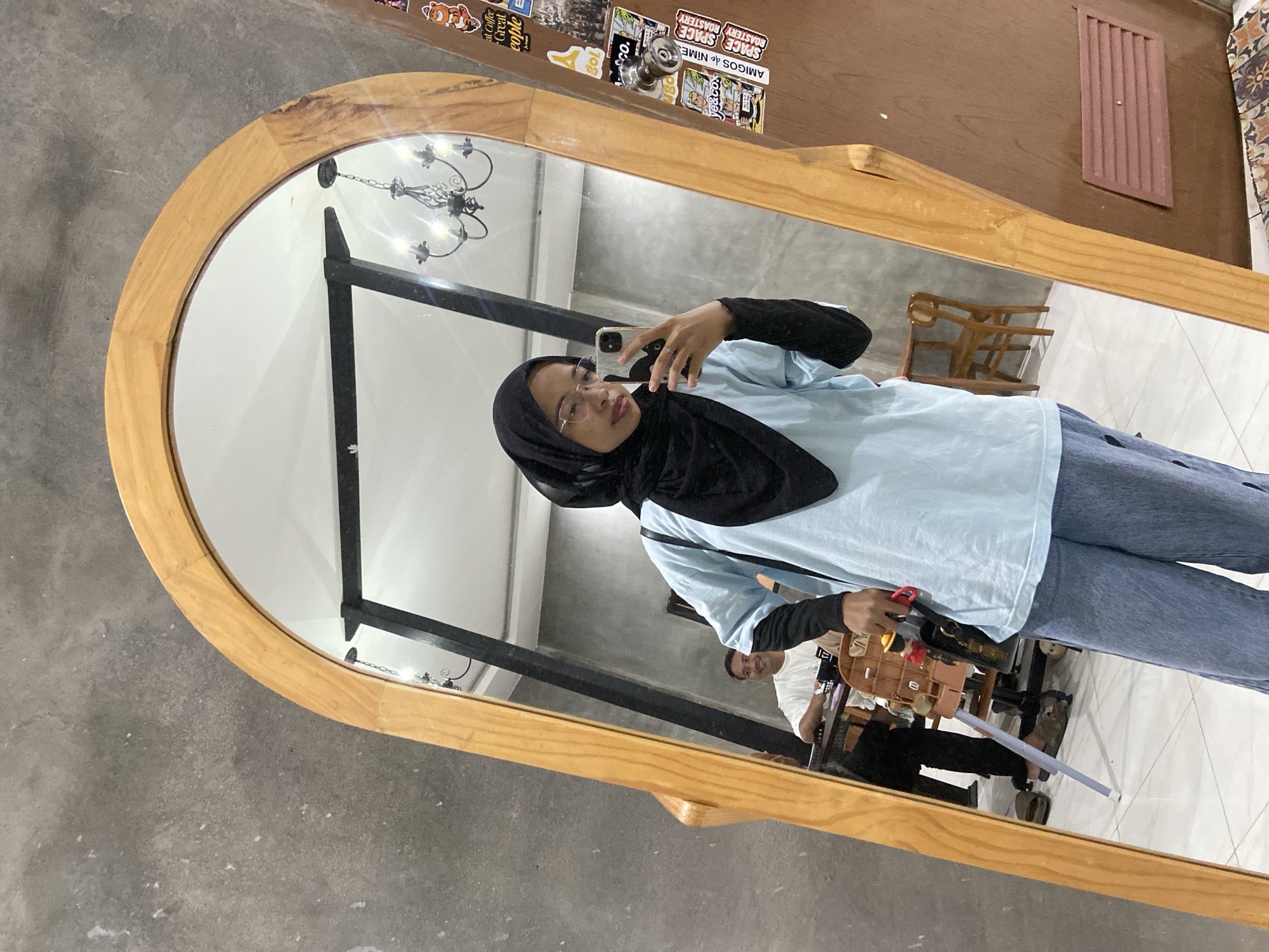 A young woman wearing a black hijab and light blue t-shirt takes a selfie in a large, arched wooden-framed mirror. She is also wearing blue jeans and glasses, and holds a smartphone in her right hand. Reflected in the mirror, a man in a white t-shirt sits at a table behind her. The background of the reflection shows a chandelier hanging from the ceiling and a concrete wall. Stickers are adhered to the right side of the mirror frame. The scene appears to be indoors, possibly in a cafe or shop, given the furniture and decor. The floor is tiled. The overall setting suggests an informal, casual environment.