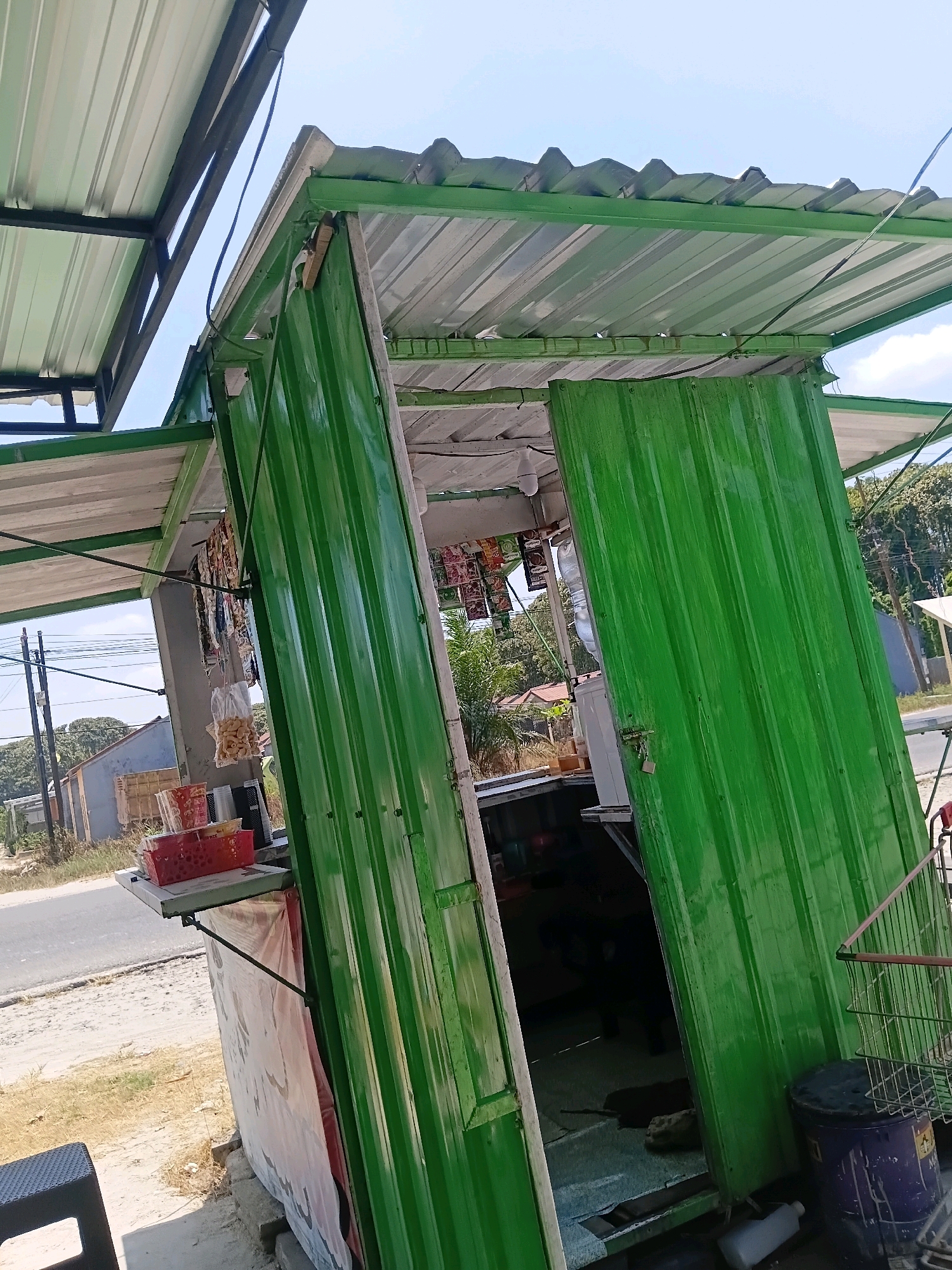 The image shows a small, open-air food stall with green corrugated metal walls and a silver corrugated metal roof supported by a metal frame. The stall is situated next to a paved road, with a dusty shoulder to its left. In the background, there are houses, trees, and power lines, suggesting a semi-urban or rural setting. The stall appears to be stocked with various snacks and beverages, indicated by packets of food hanging on display and containers on a shelf. A small padlock secures one of the stall's doors. A plastic water cooler is visible inside. To the right of the stall, a shopping cart and a large blue drum are present. The overall impression is of a modest, functional business operating outdoors.