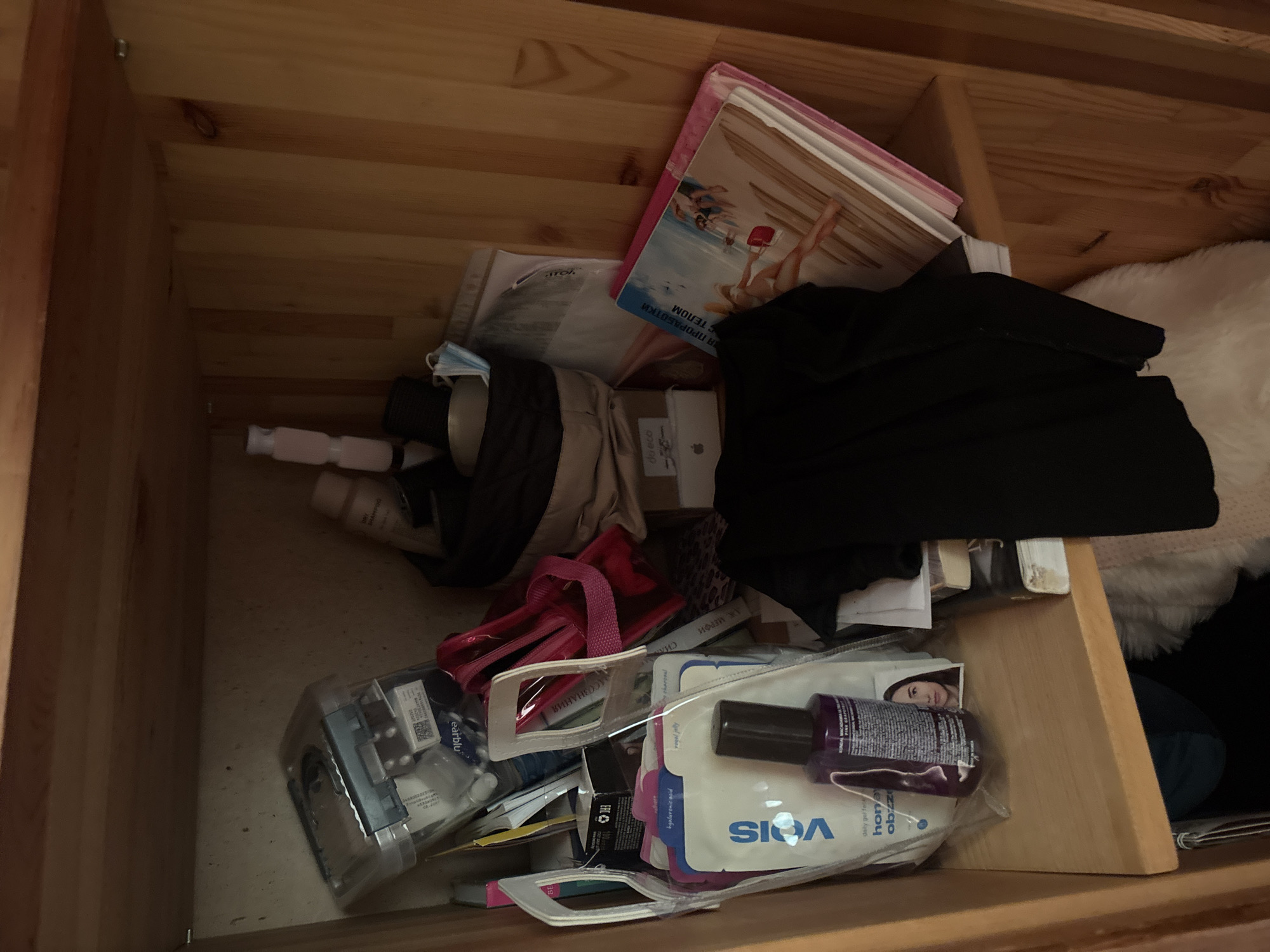 The image depicts a wooden shelf unit filled with various items, suggesting a personal storage space. On the top shelf, several bottles and containers of toiletries and cosmetics are visible, including a clear plastic case filled with smaller items, a pink cosmetic bag, and a beige pouch. On the shelf below, there are more personal care products, including a bottle of "vois" brand product with a purple liquid, and facial masks. A black fabric item, possibly clothing, is draped over some of the items on this shelf. To the right, a stack of books or magazines is leaning against the back of the unit. The overall impression is one of a cluttered but organized storage area. The location of Krasnaya Polyana, Russia, is not discernible from the visual content of the image.