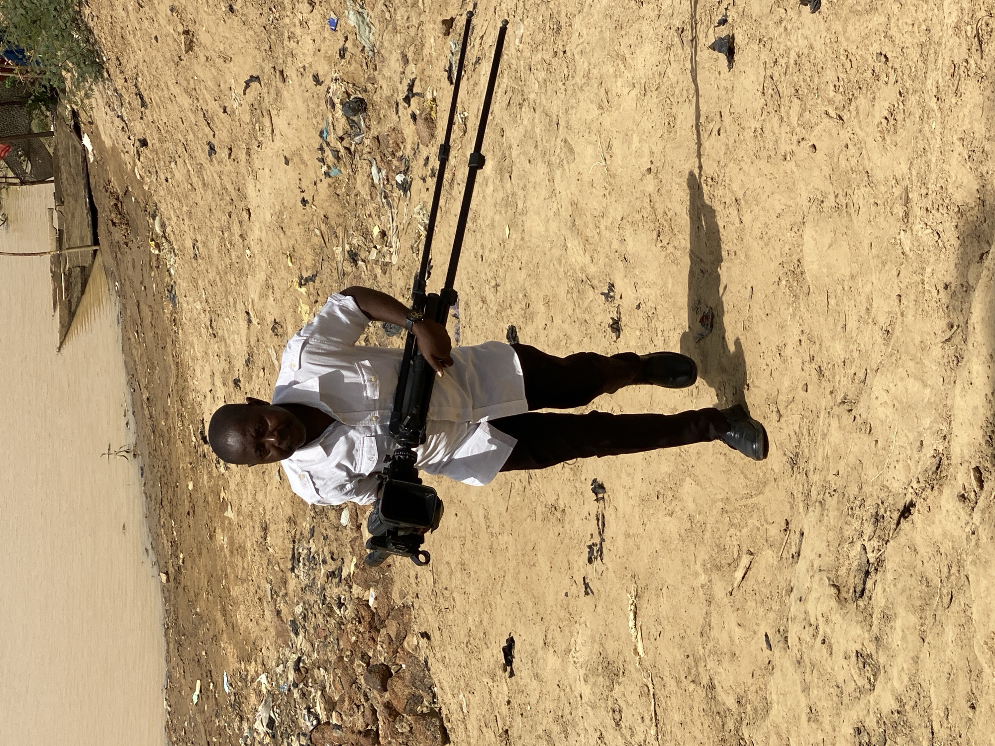 A man stands with a professional video camera on a tripod in Rigachikun, Nigeria. The man is wearing a white, button-down shirt and black pants. The terrain consists of dirt, trash, and rocks. A body of water is to his left.