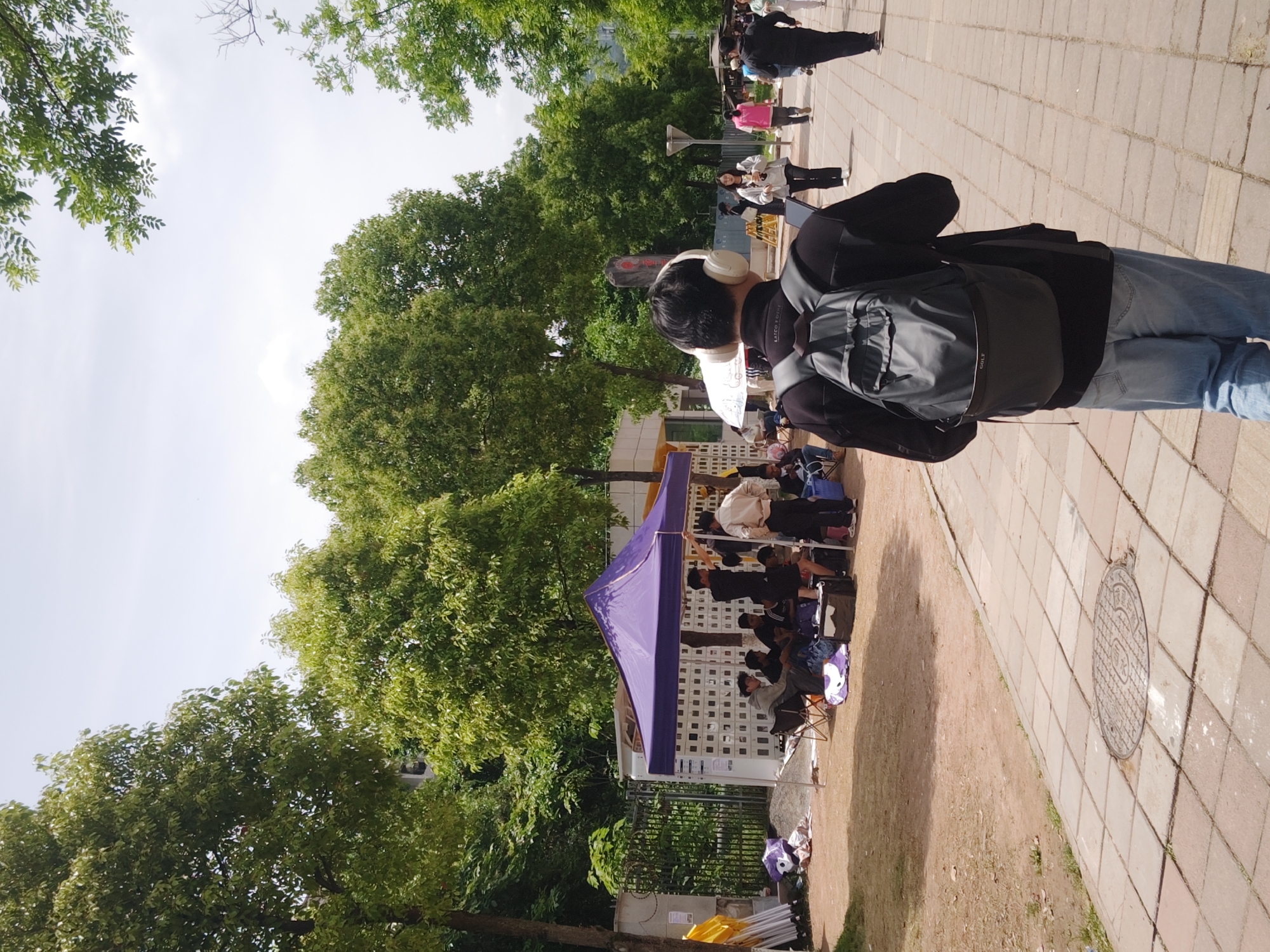 A person with headphones and a backpack is walking on a brick-paved sidewalk. To the left, there are several people sitting under a purple tent. The tent is next to a grassy area with trees and bushes. In the background, there are more people walking on the sidewalk. The scene is set outdoors in Shapingba District, China.