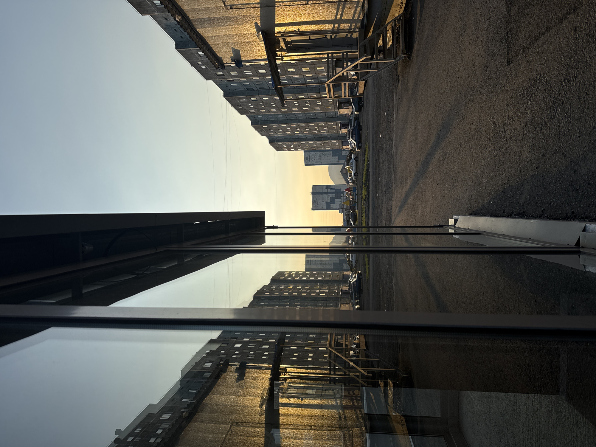 The image depicts an urban landscape at dusk, possibly in a northern city due to the soft, golden light and the architecture. The scene is divided by a large glass pane, likely from a building's storefront, which reflects the opposite side of the street.
On the left, the reflection shows a building with a golden facade, illuminated by the low sun. The building is tall and appears to be residential, with numerous windows. Metal stairs leading up to an entrance are visible in the reflection.
The actual street on the right is a paved area with several parked cars. Rows of similar residential buildings line the street, their grey facades stark against the warm sky. The sky itself is a pale blue transitioning to a soft yellow at the horizon, indicating either sunrise or sunset. Shadows are cast across the pavement, elongated by the low angle of the sun. In the distance, more buildings and what appear to be industrial structures or taller residential blocks can be seen. The overall impression is one of a functional, somewhat austere urban environment bathed in the warm glow of twilight.