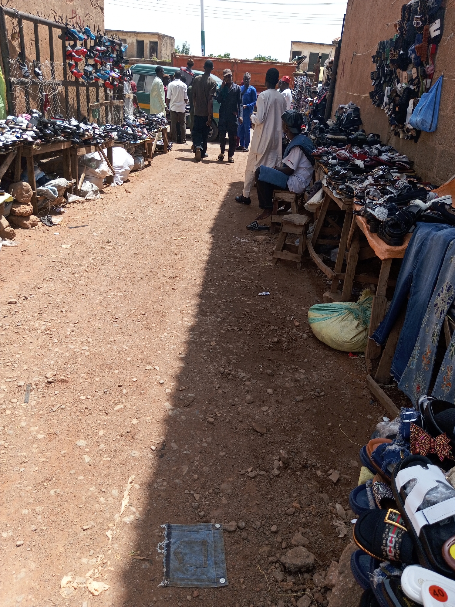 The image shows a busy street market in Bukuru, Nigeria, with a focus on a shoe stall. The ground is dusty and covered in loose soil and small stones. On the left side of the image, a stall displays numerous pairs of sandals and shoes, neatly arranged on wooden shelves and hanging from a metal rack. These footwear items vary in color and style, with some appearing new and others showing signs of wear. A few white plastic bags and some rocks are piled up near the stall.
Further down the street, several people are visible. Some are standing and browsing the merchandise, while others are walking. One man in a white traditional outfit is standing near a green van. Another man in a dark green shirt and dark trousers is walking away from the camera. A woman in a blue hijab and white top is seated on a wooden stool in front of a stall laden with more shoes, which are piled high on a wooden table.
On the right side of the image, another stall displays more footwear, along with some hanging denim jeans. A large, light green tarp is spread on the ground near this stall. The wall of a building forms the backdrop on the right, with its rough, plastered surface and some electrical wires overhead. The overall scene depicts a vibrant marketplace with a focus on the sale of footwear, characteristic of urban street commerce in Nigeria.
