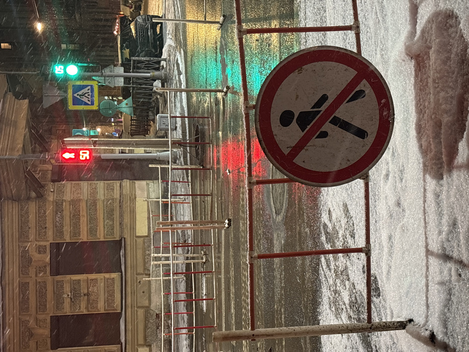 A nighttime winter scene depicts a city street with slush and snow. A "No Falling" sign stands to the right, attached to a temporary barrier. Traffic lights show red to the left and green further ahead. A pedestrian crossing sign is also visible. A building with patterned walls flanks the left. Snow is visible on the right.