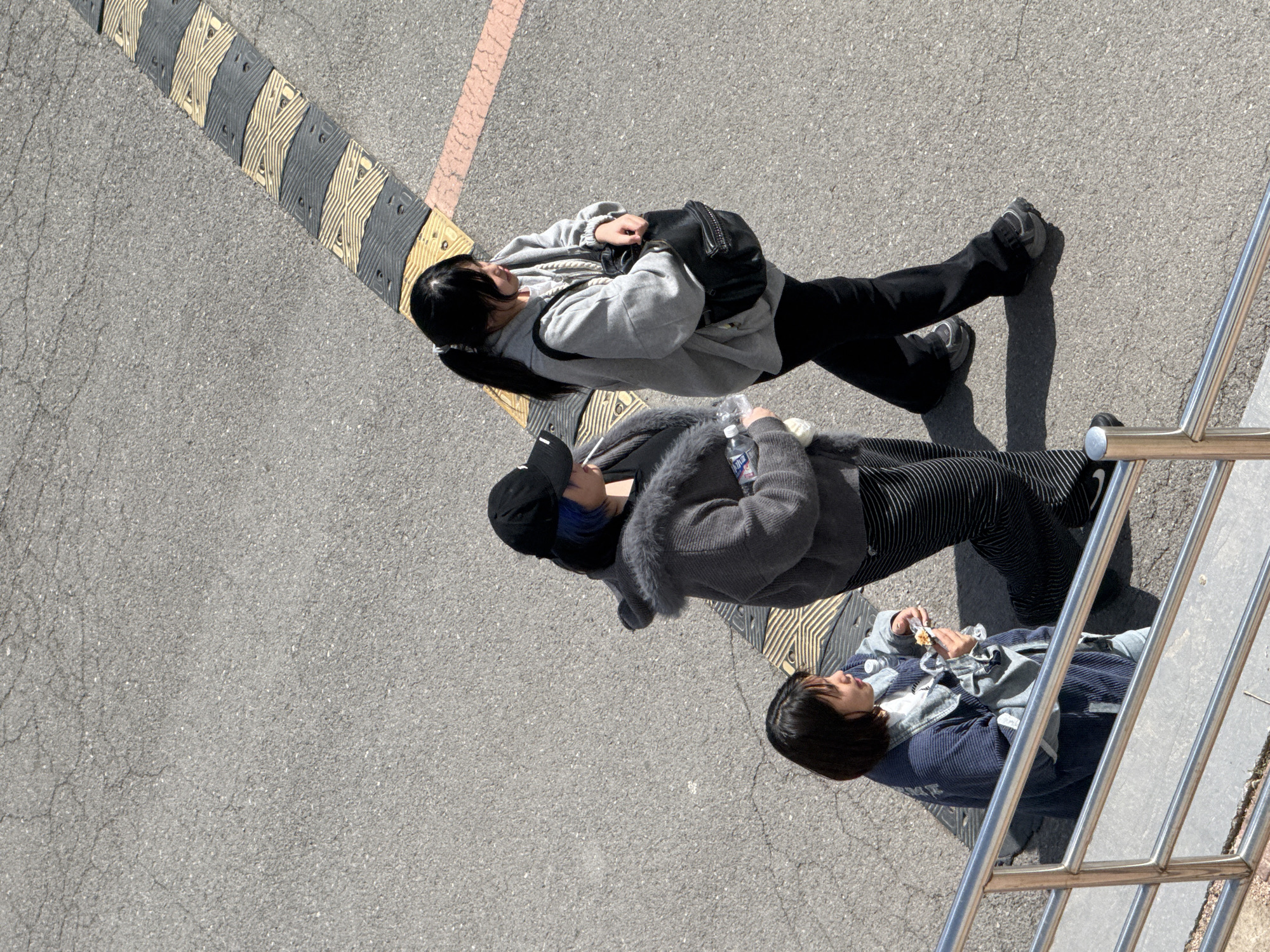 The image, taken from an elevated perspective, depicts three people on a paved surface in Zhongtun, China. The individuals are positioned near a speed bump marked with yellow and black stripes and a red line.
The first person, at the top, wears a light gray hooded sweatshirt, black pants, and dark sneakers. They have a black bag slung across their body.
The second person wears a dark baseball cap, a gray coat with a fur collar, and striped pants. They are holding a plastic water bottle.
The third person, nearest the metal railing on the right, wears a blue and gray jacket. They appear to be holding something in their hands.
The ground is asphalt with patches. A metal railing is visible in the lower right corner.