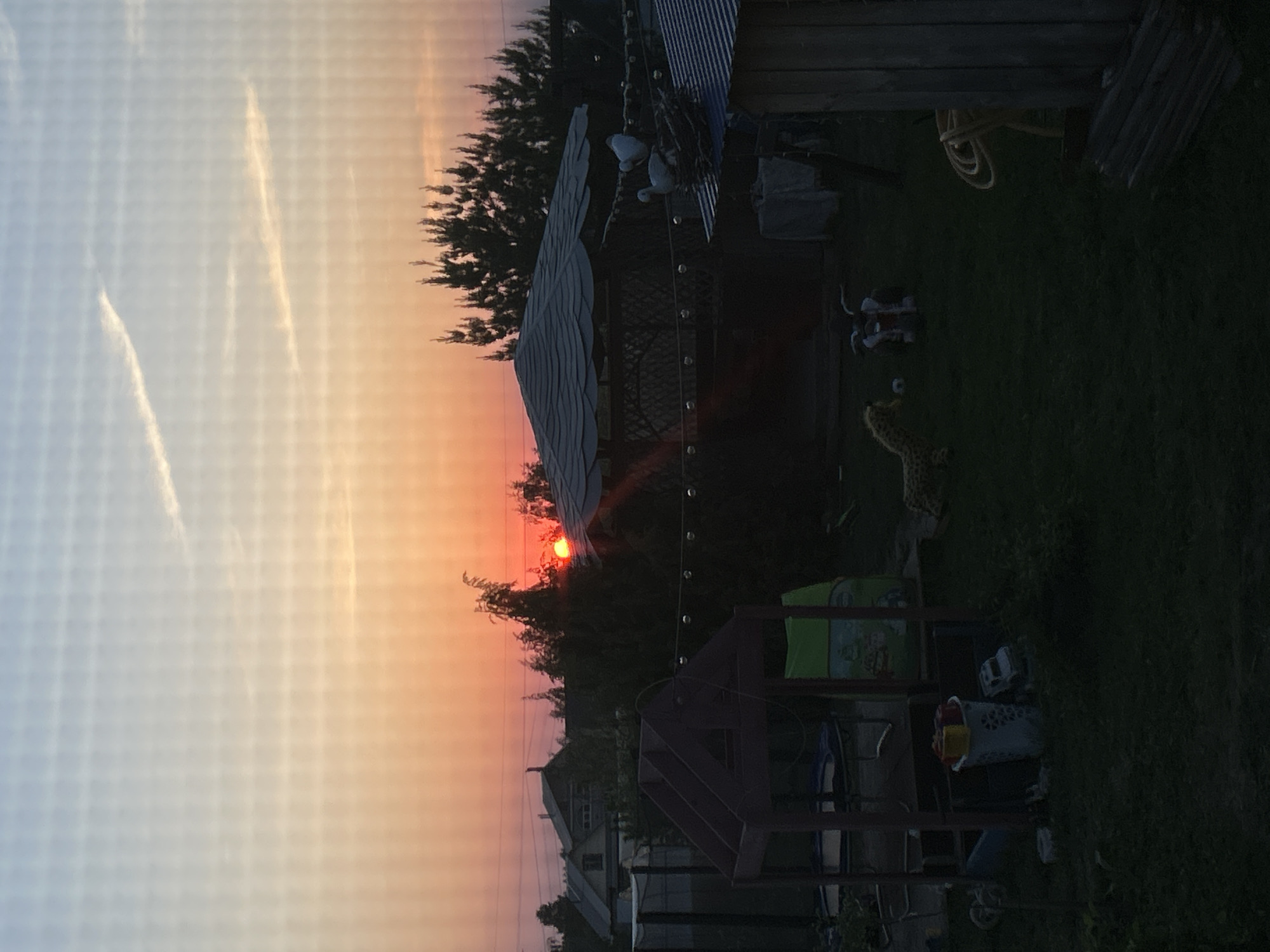 The image depicts a backyard scene at sunset in Ольховка, Russia. A red-orange sun is partially obscured by trees, casting an orange hue across the sky. In the foreground, a children's playset is visible. A gazebo with a corrugated metal roof and hanging lights is partially silhouetted against the setting sun. A few toys, including a stuffed animal, are scattered on the grass. In the background, houses are discernible in the dim light. The sky shows contrails.