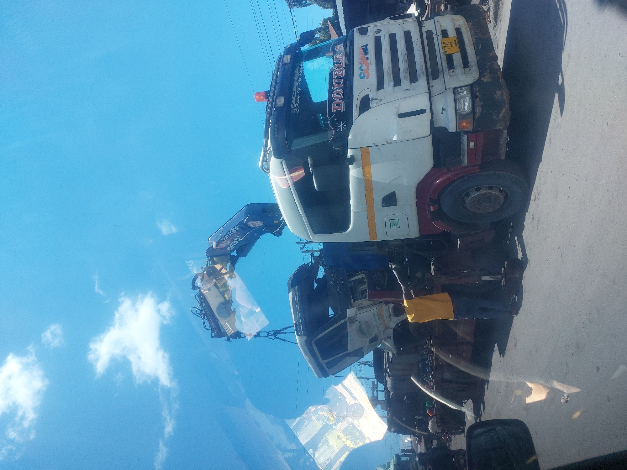 The image, viewed upright, depicts an outdoor scene under a bright, clear blue sky with scattered white clouds. The primary subject is a large white and maroon Scania truck, equipped with a crane, parked on a paved surface.
The Scania truck's cab is white with maroon accents, and the front grille is somewhat weathered. On the windshield banner, the words "DOUBLE S" and "SCANIA" are prominently displayed, along with "King Of The Road". A yellow license plate on the front bumper is partially visible, showing "T 740 DDL". An orange rotating light is mounted on the cab's roof.
Attached to the Scania truck is a hydraulic crane, which is actively engaged in lifting or holding what appears to be another vehicle's chassis or a significant component of a vehicle. The object being lifted is light-colored and somewhat rectangular. Several individuals are present around the vehicles. One person in a yellow shirt and blue trousers is visible near the ground next to the Scania truck. Others are partially obscured by reflections or are further in the background, observing or assisting with the operation.
The scene suggests a recovery, repair, or loading activity. Strong shadows are cast on the ground, indicating bright daylight conditions. Power lines are visible stretching across the upper background. The foreground features significant reflections on what appears to be a car window, showing distorted views of the sky, clouds, and parts of the street and vehicles. The general setting is consistent with a roadside or service area in Dar es-Salaam, Tanzania.