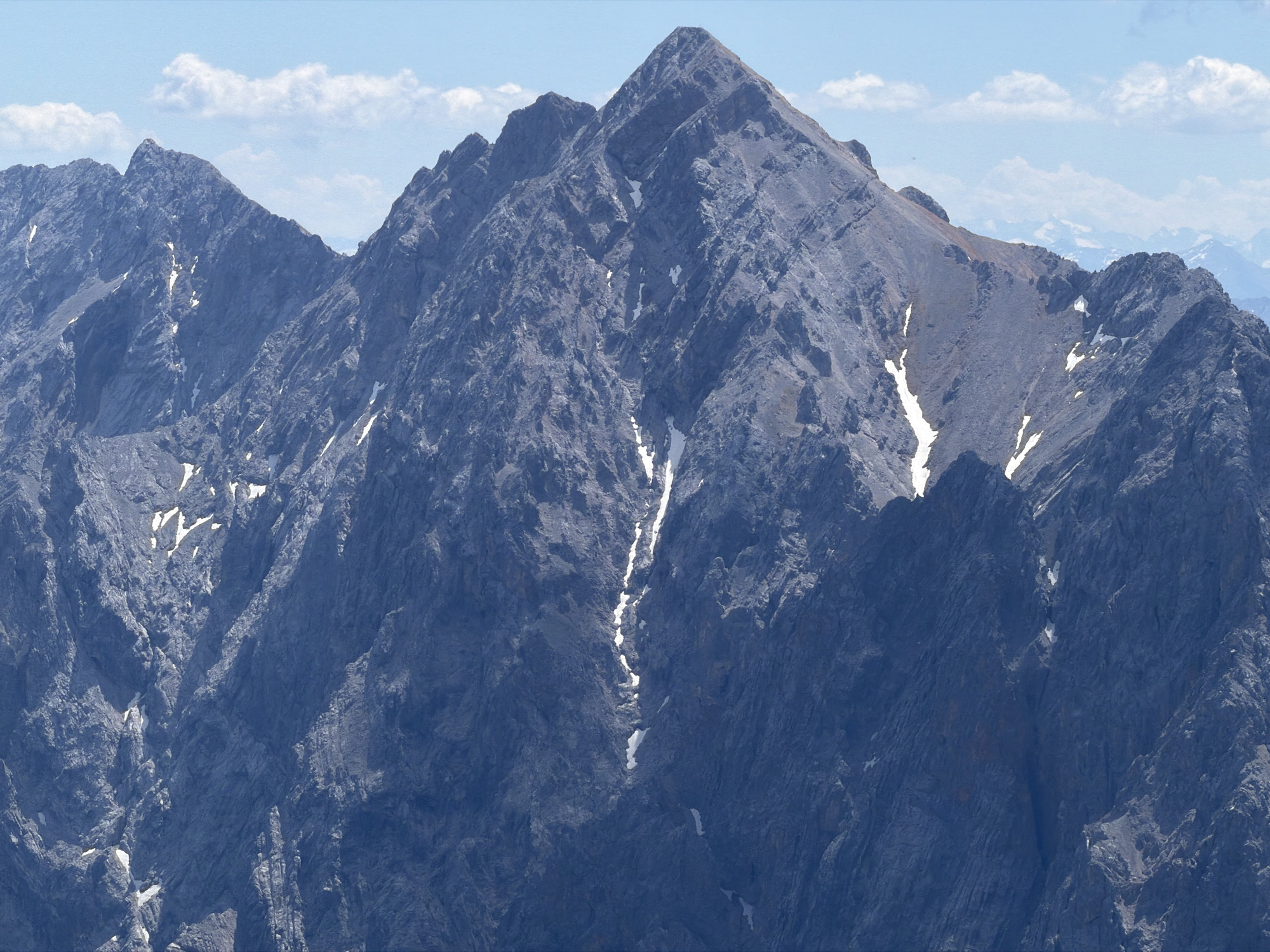 This is a medium-range, eye-level, full shot photograph of the Zugspitze, the highest peak in Germany, located near Garmisch-Partenkirchen. The mountain range dominates the frame, with its rugged, rocky slopes rendered in deep blues and grays. Patches of white snow are visible in crevices and on some of the higher ledges, contrasting with the dark rock. The sky above is a pale blue with a few wispy white clouds. The overall impression is one of imposing natural grandeur and a sense of high altitude. There are no people or man-made structures visible in this particular frame.