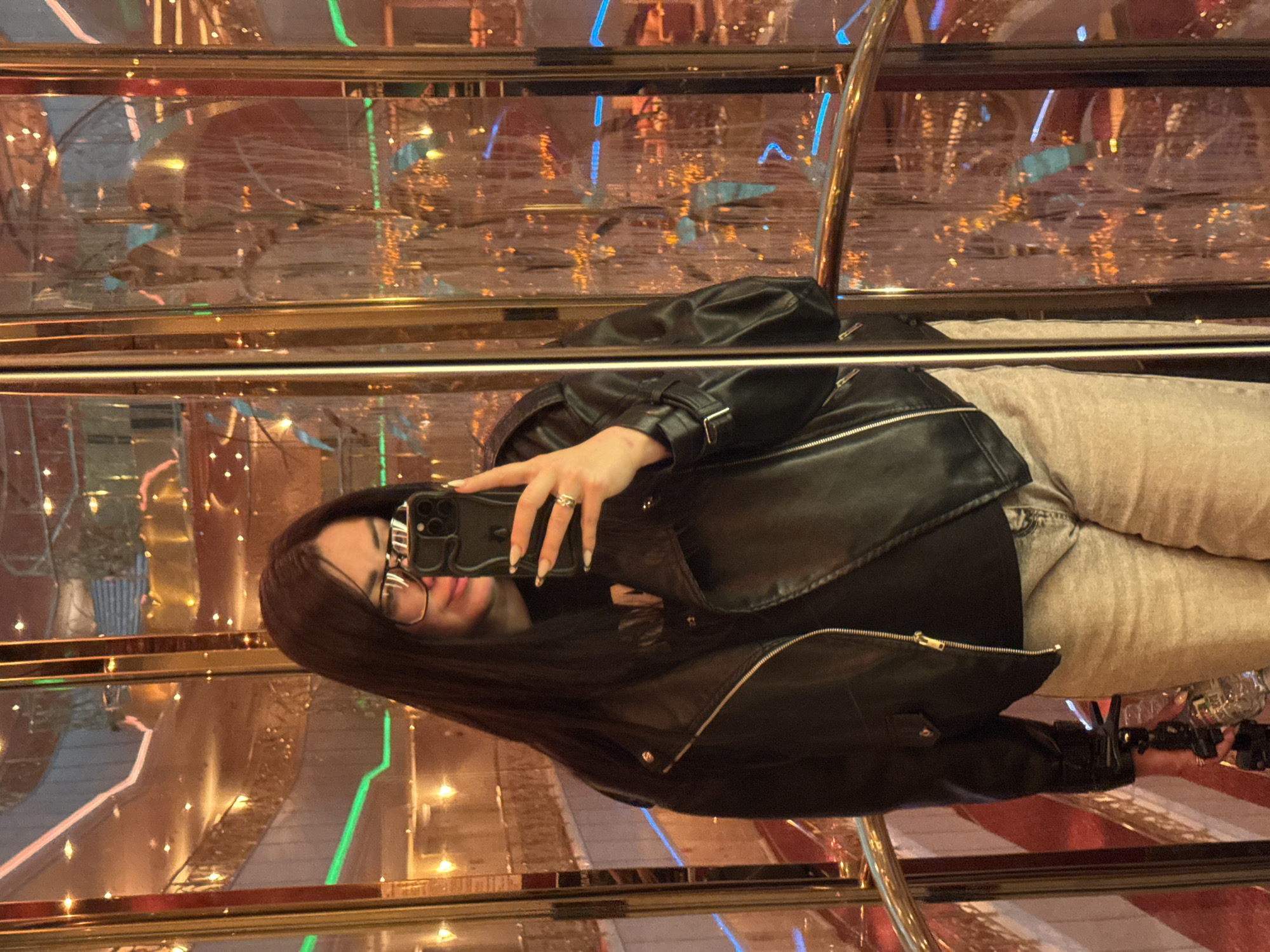 The image displays a person taking a selfie within a highly reflective and illuminated indoor environment. The subject, a person with long, dark hair and glasses, is seen wearing a black leather jacket with zippers and buttons, paired with light-colored pants. They are holding a dark-colored smartphone, likely an iPhone given its camera array, in their right hand to capture the photo. A ring is visible on the ring finger of the right hand, and the nails appear manicured with a light tip.
The setting is characterized by extensive use of reflective surfaces, including polished golden or brass-toned railings and mirrored panels, which create a complex, multi-dimensional backdrop of reflections. Numerous bright lights, primarily warm yellow, orange, and red, are interspersed with cooler blue and green lights, indicating an artificially lit interior. This ambiance suggests an evening or nighttime scene within an ornate or upscale venue such as a hotel lobby, a cruise ship, or an entertainment complex. Faint outlines of a bottle and other indistinct objects are visible in the background, obscured by reflections. There is no visible text in the image.