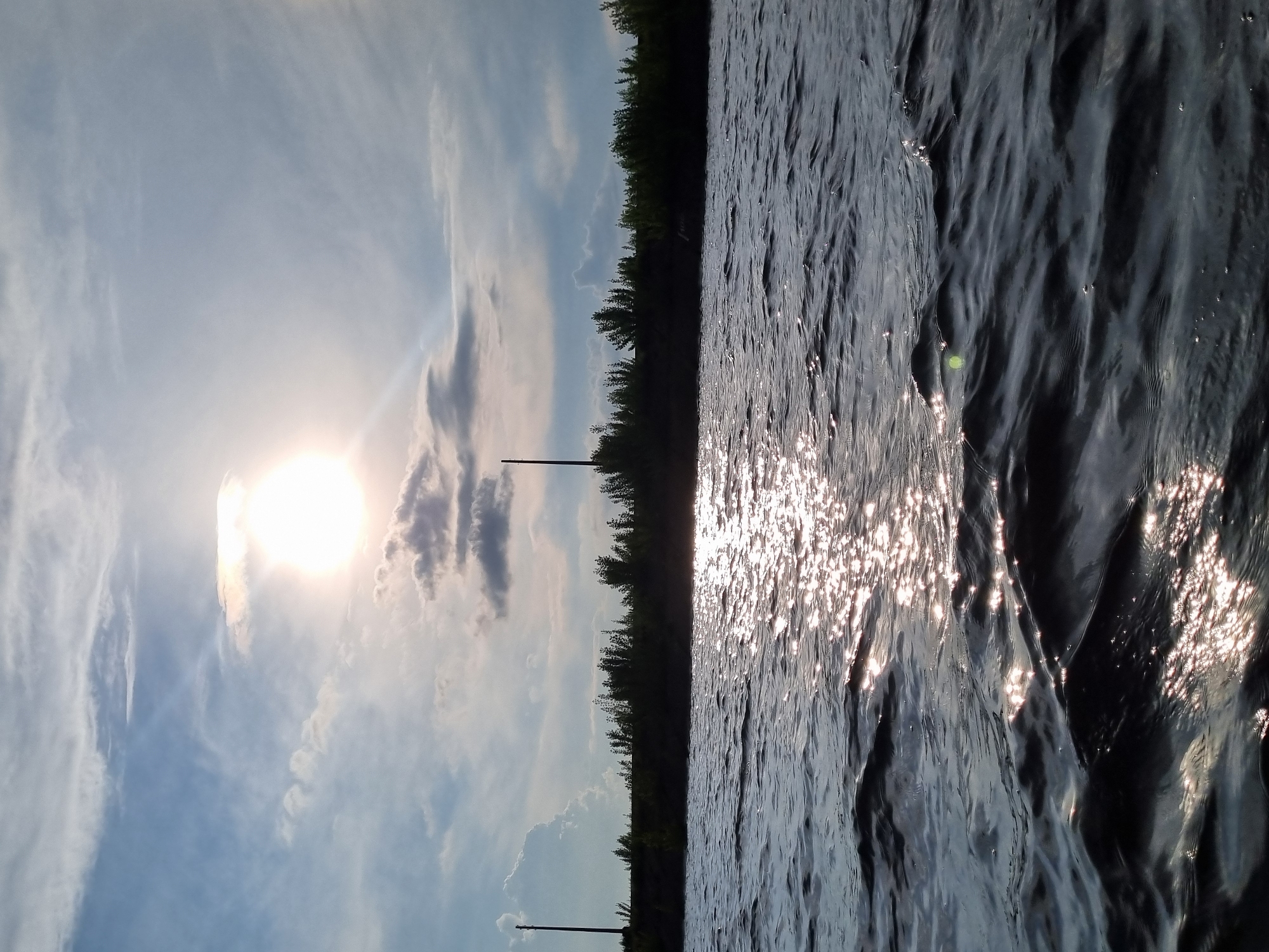 The image captures a serene river landscape under a partly cloudy sky with the sun partially obscured by clouds. The foreground is dominated by rippling water, reflecting the sunlight in shimmering streaks. In the middle ground, a dark, silhouetted tree line forms the horizon, punctuated by two tall, thin poles, one on the left and another further to the right, likely power line poles. The overall impression is one of a tranquil natural scene, possibly taken from a boat or the riverbank. This location appears to be the Kama River near Chaikovsky, Russia, given the characteristic power lines along the river and the dense vegetation often found in that region.
