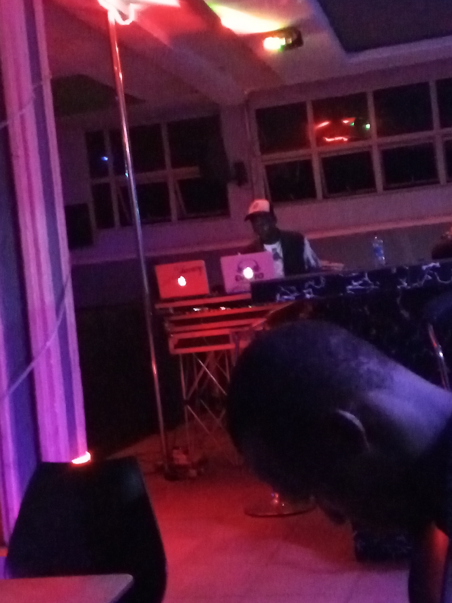 The image shows an indoor scene, possibly a club or bar in Lokoja, Nigeria, illuminated with red and purple lighting. In the background, a man wearing a cap and dark clothing stands behind a DJ setup consisting of two laptops (one with an Apple logo and another with a pink logo), mixers, and equipment on a raised platform. A stripper pole is visible to the left of the DJ booth. In the foreground, the top of a dark-haired man's head and part of his face are visible. A black speaker sits in the left foreground. Windows can be seen in the background, indicating an indoor space. A water bottle is on the DJ’s table.