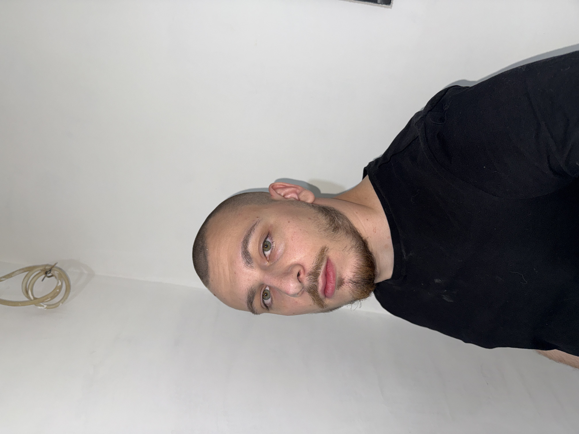 The image displays a head-and-shoulders shot of a young man, rotated 90 degrees counter-clockwise, against a plain white wall. He has a closely-shaven head, a dark beard and mustache, and prominent green eyes. The man looks directly at the camera with a neutral to slightly serious expression. He is wearing a black t-shirt, which appears to have some light smudges or dust. In the upper left portion of the background, a coiled, light-colored cable or tube is visible, attached to the white wall. The setting is an indoor space, characterized by its stark, unadorned white walls, suggesting a simple or potentially unfinished room. The lighting is bright and even. No text is visible in the image. The location context for this media file is Stara Zagora, Bulgaria.