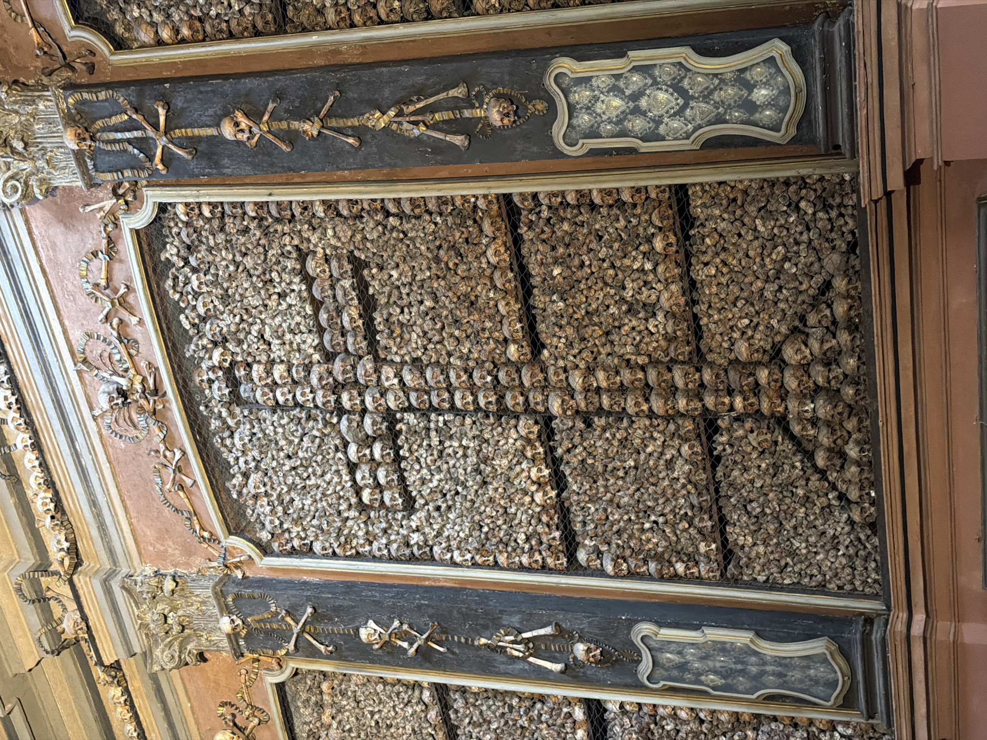 The image depicts the ossuary of San Bernardino alle Ossa, a church located in Milan, Italy. The walls and ceiling are adorned with human skulls and bones, arranged in decorative patterns. The bones appear aged and weathered, ranging in color from beige to brown. The ossuary is divided into panels framed by dark, ornate molding. Within the panels, the bones are packed tightly together, forming dense, textured surfaces. Skulls are prominently featured, often arranged in rows or patterns. Interspersed among the skulls and bones are decorative elements, including skeletal figures and crests. The overall effect is both macabre and artistically elaborate.