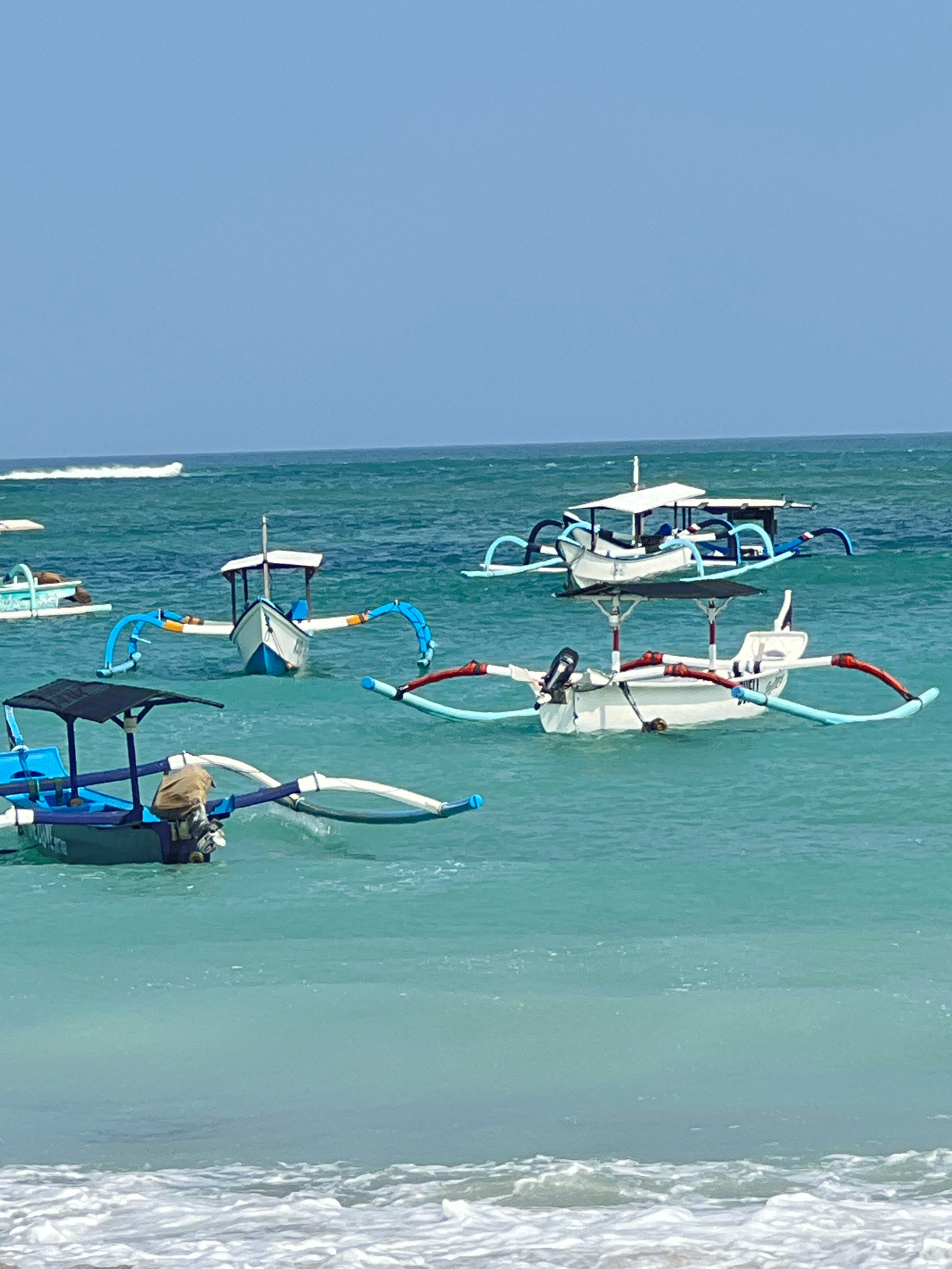 The image depicts a serene beach scene in Kuta, Indonesia, with several traditional Balinese fishing boats, known as outrigger boats, floating on the turquoise waters of the Indian Ocean. The boats are predominantly white and blue, with outrigger systems that provide stability. Some boats have canopies for shade, and one boat in the foreground has a person sitting in it, draped in a tan cloth. Another boat behind it has an outboard motor. The water is clear, with gentle waves lapping at the shore in the foreground. The sky above is a clear, pale blue, indicating a sunny day. The composition suggests a peaceful moment at a popular tourist destination.