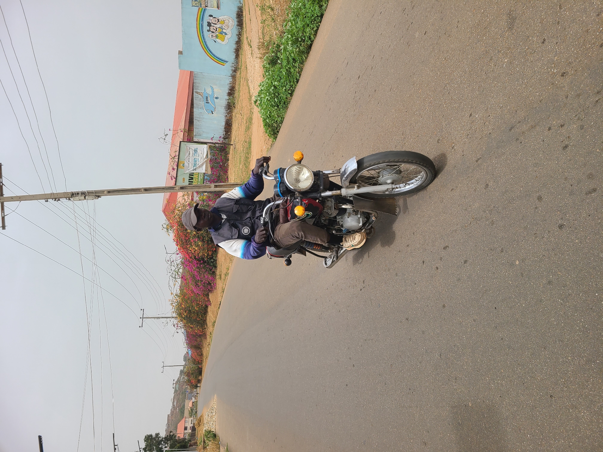 A man is riding a motorcycle on a paved road. He is wearing dark clothing and gloves. The road is bordered by vegetation on one side. Power lines run along the road. Buildings and a mural are visible in the background. The location is Barakin Lerawi, Nigeria.
