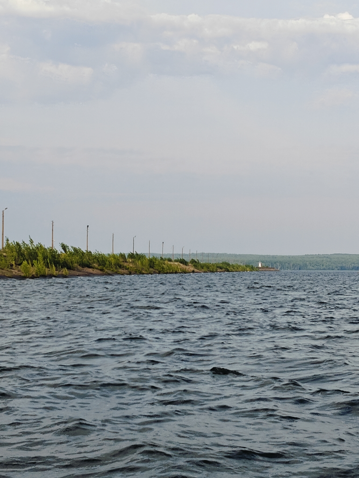 A body of dark blue water stretches across the foreground of the image, its surface rippled by a gentle breeze. In the background, a line of green trees and bushes forms a dense border along a sandy embankment. A series of tall, thin poles, some with lights, are spaced out along the edge of the land. In the distance, a white lighthouse stands on a small island. The sky above is a pale blue, with scattered white clouds. The scene appears to be the Kama River in Chaikovsky, Russia.