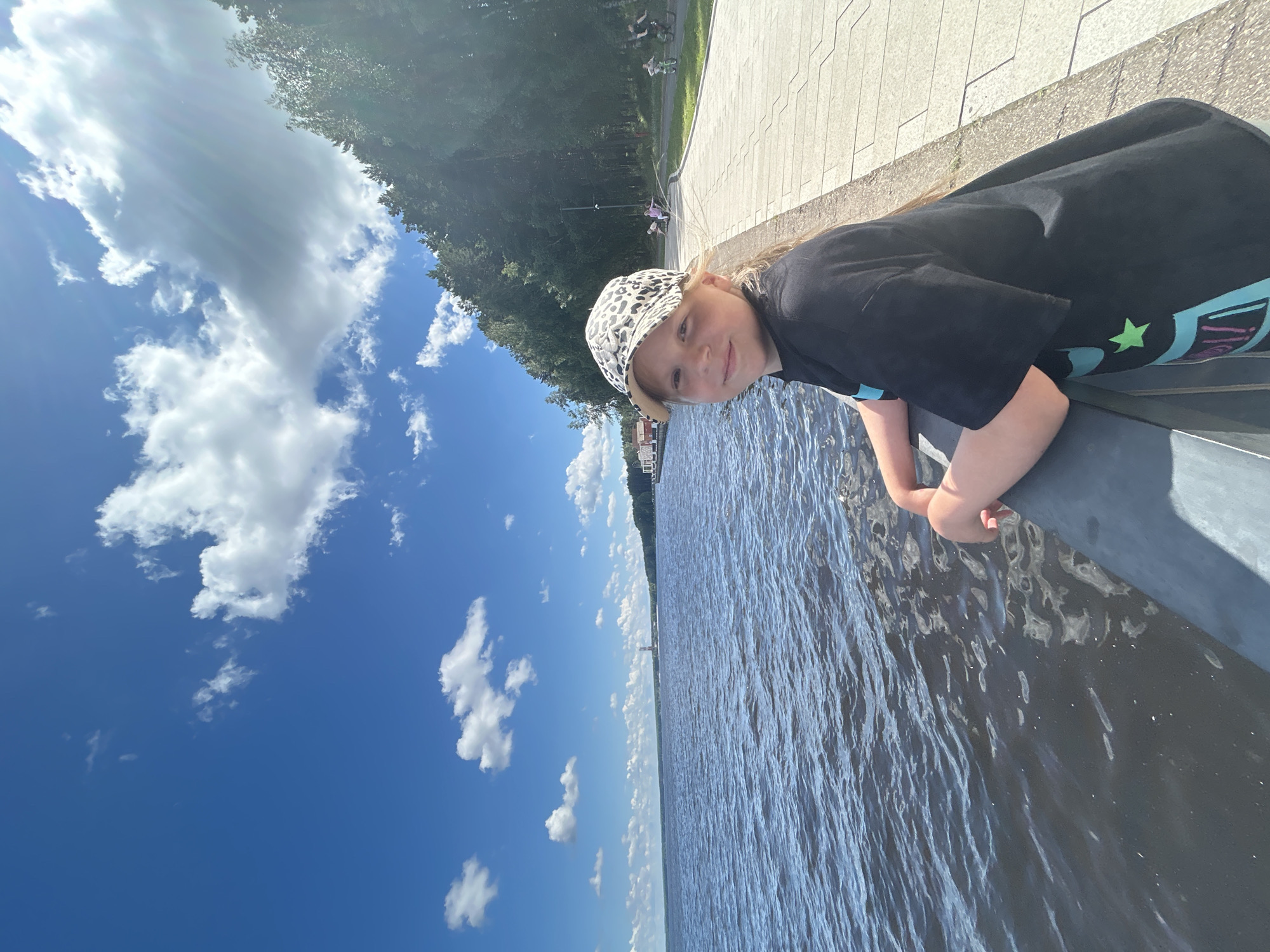 The image features a young girl wearing a black t-shirt with a colorful graphic and a leopard-print baseball cap. She is leaning on a dark-colored railing next to a body of water. Her expression is a slight smile.
The scene appears to be outdoors, possibly at a waterfront promenade or park. In the background, there is a paved walkway lined with trees and some people can be seen in the distance, including some on bicycles. The sky is a bright blue with scattered white clouds, suggesting it is daytime and the weather is clear and sunny. The water has a rippled texture, indicating a slight breeze. A faint structure, possibly a lighthouse or a pier, is visible in the distance across the water.
The image is oriented sideways, with the sky and water running vertically. There is no visible text in the image. The overall mood is cheerful and summery.