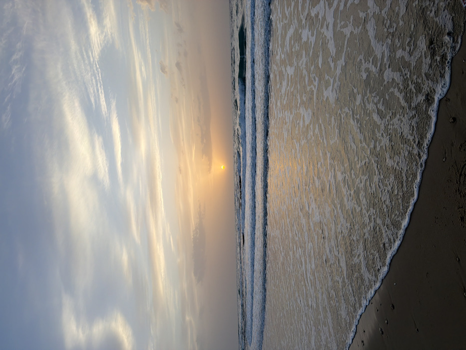 The scene depicts a sunset viewed from a beach in Tel-Aviv, Israel, facing west over the Mediterranean Sea. The sun, a distinct orange-yellow disc, is positioned low on the horizon, slightly left of the vertical center. The sky above presents a gradient from pale blue at the top, transitioning to orange and yellow hues closer to the horizon, with scattered stratocumulus and cirrus clouds. Multiple parallel waves are visible in the midground, cresting and receding, displaying white foam. The foreground consists of dark, wet sand with a clear foamy waterline marking the most recent wave recession. No individuals or man-made objects are present within the frame.