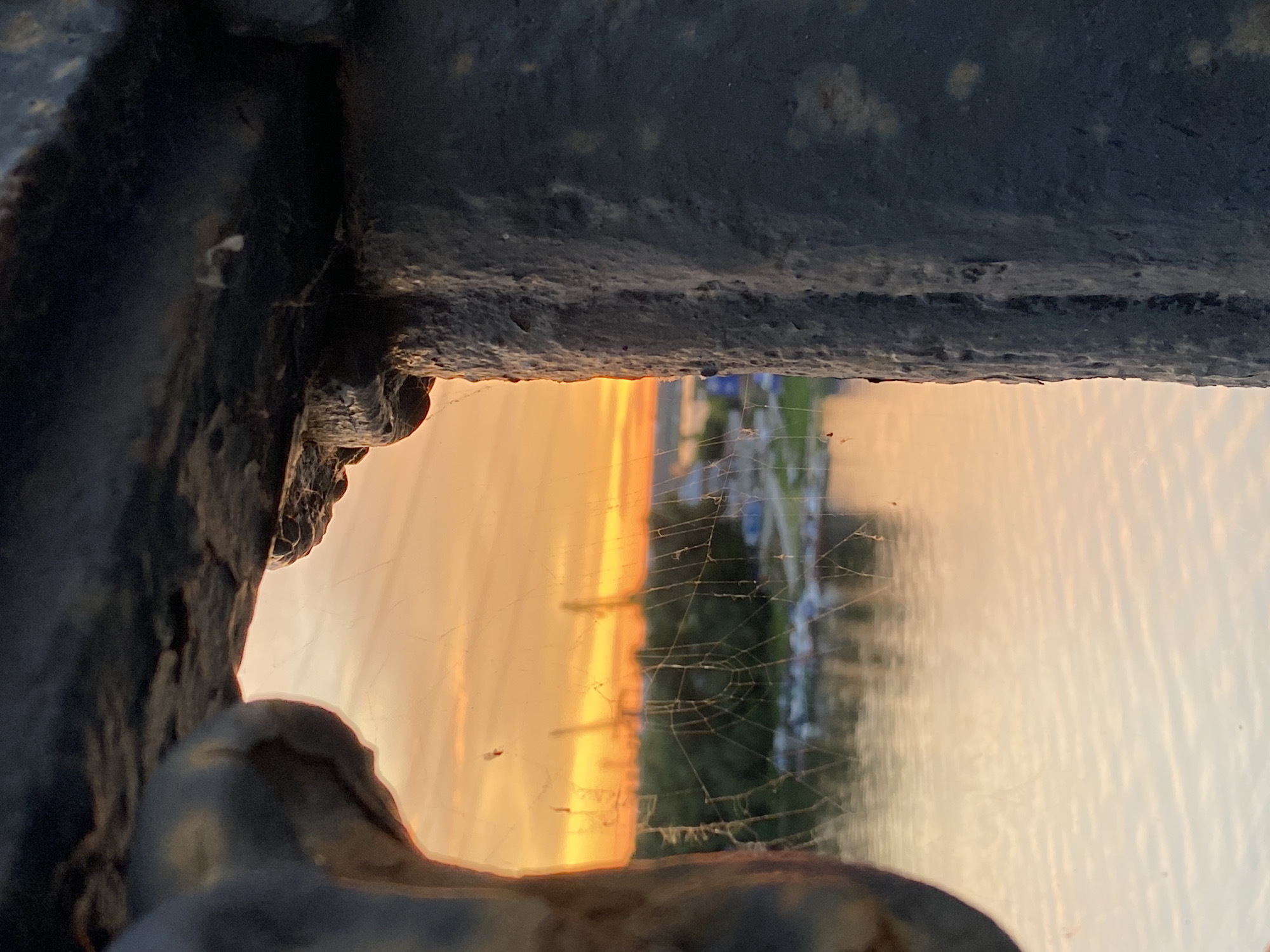 The image is a close-up, low-angle shot of a dark, weathered metallic structure, possibly a railing or a gate, with a bright, hazy sunset visible through an opening. The metallic structure dominates the foreground and the top of the frame, showing a rough, textured surface with some rust and debris. Through the gap in the metal, a vibrant sunset casts an orange and yellow glow. A thin, intricate spiderweb stretches across a portion of the opening, partially obscuring the view of the scene beyond.
The background scene is out of focus but appears to be a body of water, possibly a river or lake, reflecting the colors of the sunset. Hints of green foliage and some indistinct structures, perhaps buildings or boats, can be seen on the far side of the water, suggesting an outdoor location. The overall impression is one of a secluded or framed view of a natural phenomenon. The time of day is clearly sunset, indicated by the warm, saturated colors of the sky. The weather appears calm, with the water surface showing gentle ripples. There are no visible people or text in the image. The emotional tone could be interpreted as peaceful, introspective, or even slightly melancholic due to the muted colors and the presence of the spiderweb. The setting appears to be near a body of water at dusk.