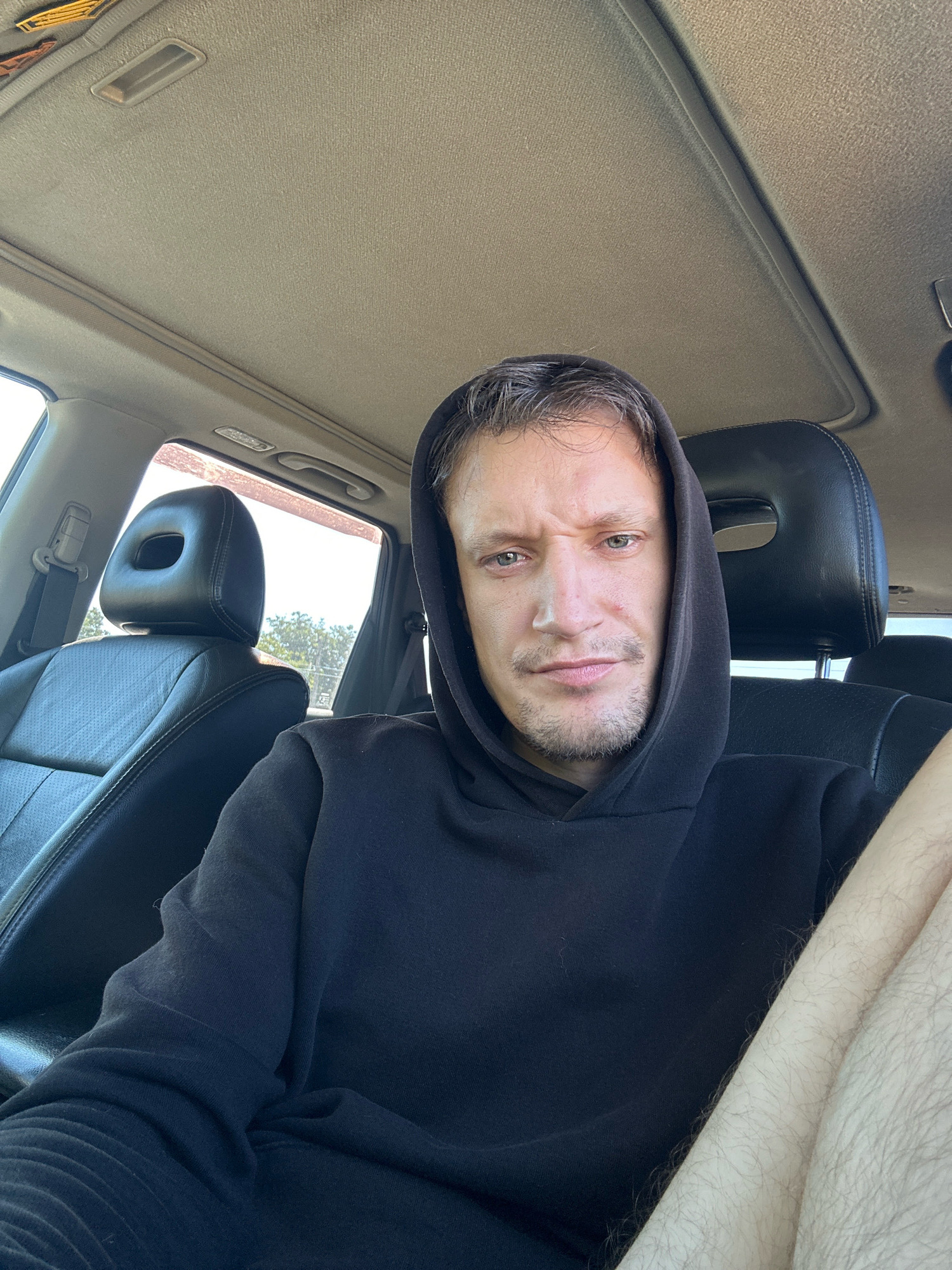 A man wearing a black hoodie sits in the driver's seat of a car, looking towards the camera. The car's interior is visible, including the headrest of the passenger seat, the dashboard, and the sunroof. The man has short, dark hair and stubble on his face, and his expression appears neutral. The setting is daytime, with sunlight streaming into the car. The image is a selfie taken from the man's perspective. The location is the city of Makiv, Ukraine.