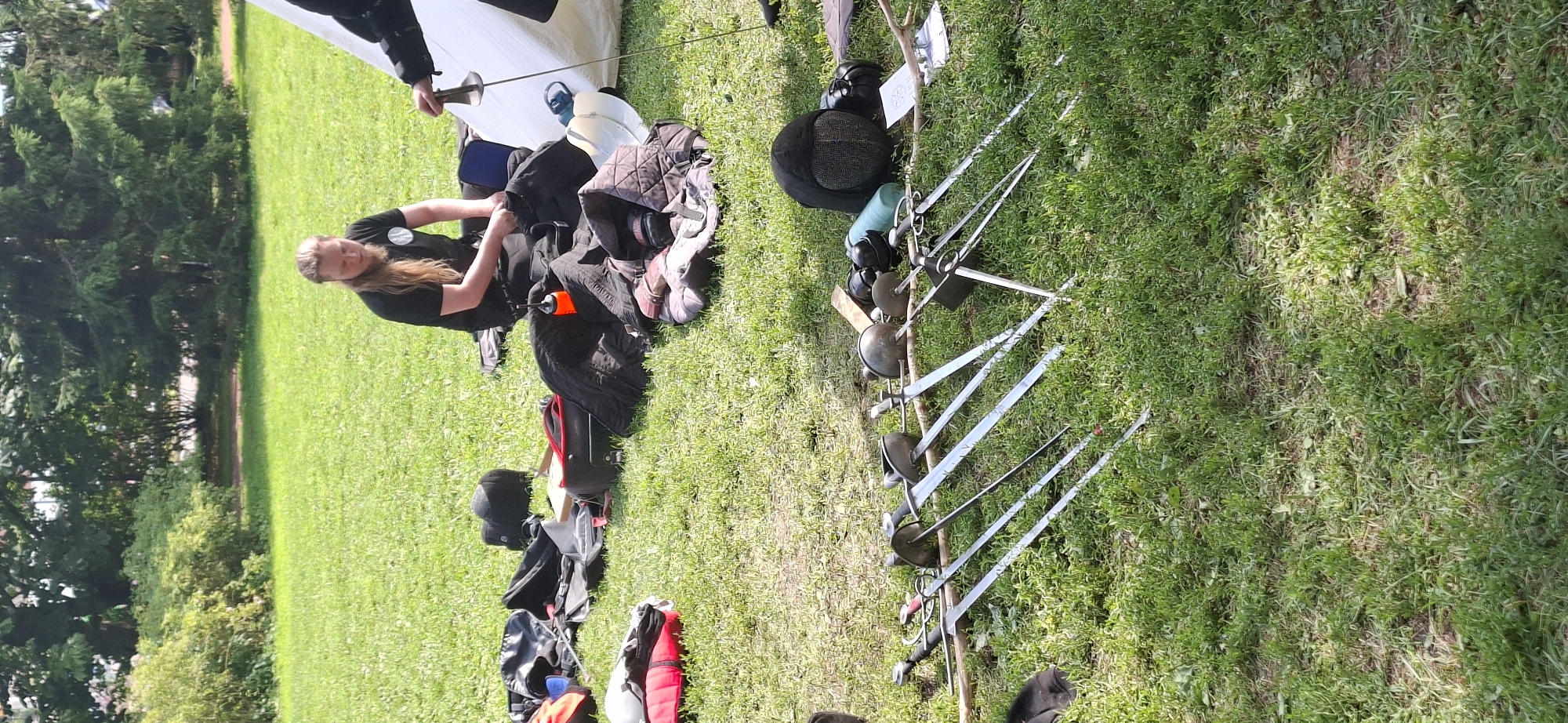 A group of people are engaged in a historical fencing practice session in a park, likely in Turku, Finland, given the provided context. In the foreground, several fencing foils and sabers are laid out on the grass, arranged in a line. These weapons appear to be part of the equipment for the practice. To the right, a person dressed in fencing attire, including a mask and gloves, is holding a foil. Behind the swords, there are various bags, mats, and protective gear scattered on the grassy ground, indicating preparation or aftermath of the activity. In the middle ground, a person with long blonde hair, wearing a black t-shirt, is seated on the grass, looking down at something in their hands, possibly adjusting equipment. The environment is a sunlit park with lush green grass and trees in the background, suggesting an outdoor recreational area.