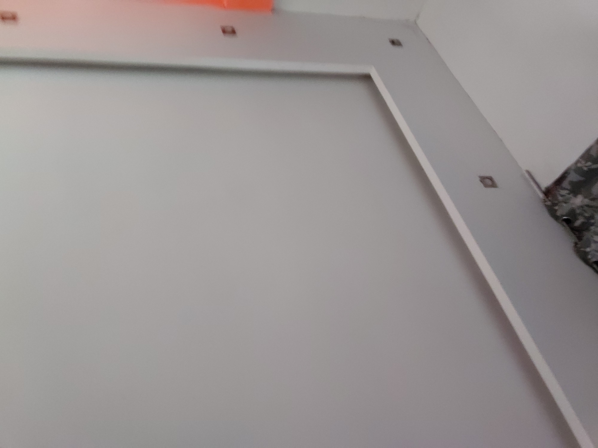 The image presents an indoor scene within a building in Santa Ana, Venezuela. The primary subject is a large, light gray wall or panel, possibly a door, bordered by a distinct white trim that creates a clean, rectangular outline. Above this trim, on the light gray wall, several small, dark, square objects are affixed at regular intervals, which appear to be either electrical box covers or mounting points for fixtures.
At the very top left of the frame, a blurred orange object is partially visible. To the right, a white wall meets the gray wall, forming an internal corner of the room. On the far right edge of the image, a small portion of a dark, patterned fabric, possibly from a curtain or upholstery, can be seen. The overall impression is of a clean, minimalist interior space with smooth, painted surfaces. There are no people visible, and no specific activity is taking place. The lighting is even, suggesting ambient indoor illumination. No text is visible in the image.