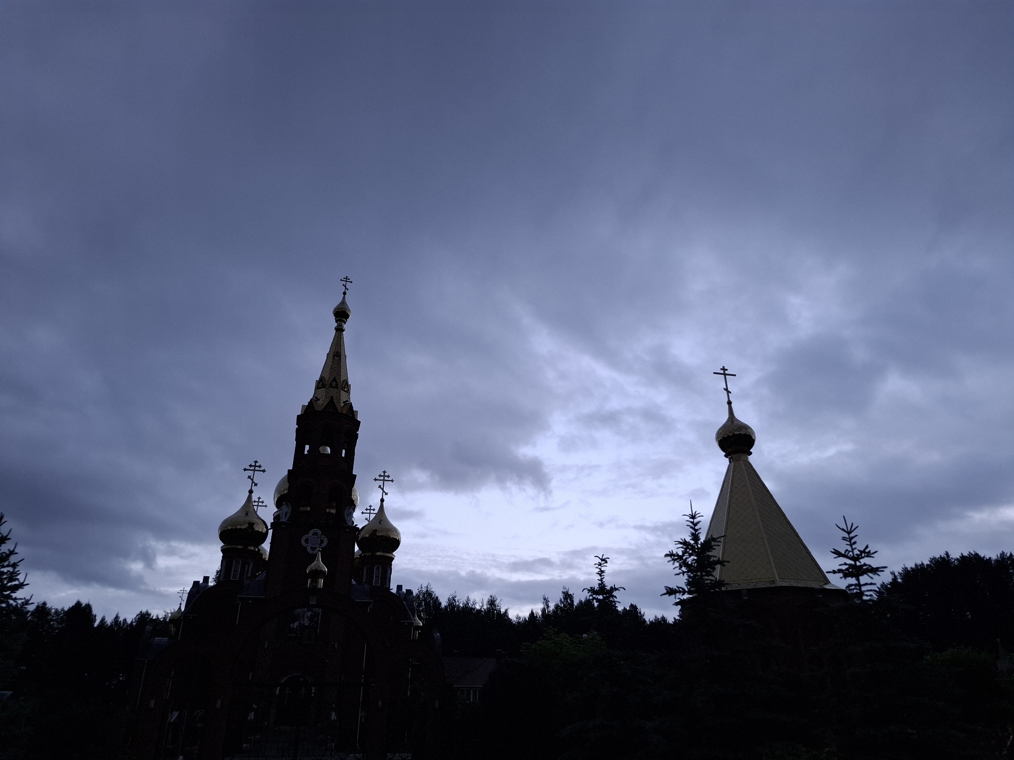 The image captures a silhouette of an Orthodox Christian church complex in the city of Chaikovsky, Russia, against a heavily overcast, dark blue to purplish-gray sky. The scene is characterized by low light, rendering most foreground and midground elements in deep shadow. In the left half, a large, multi-domed church structure is visible, featuring a prominent tall spire and several smaller onion domes, all adorned with golden Orthodox crosses. To the right, a separate, smaller structure with a golden conical roof and a single golden onion dome, also topped with an Orthodox cross, stands partially obscured by foliage. The lower portion of the image is dominated by dark, dense silhouettes of evergreen and deciduous trees, forming a natural barrier that obscures the bases of the religious structures. No people, animals, or specific actions are discernible within the frame.