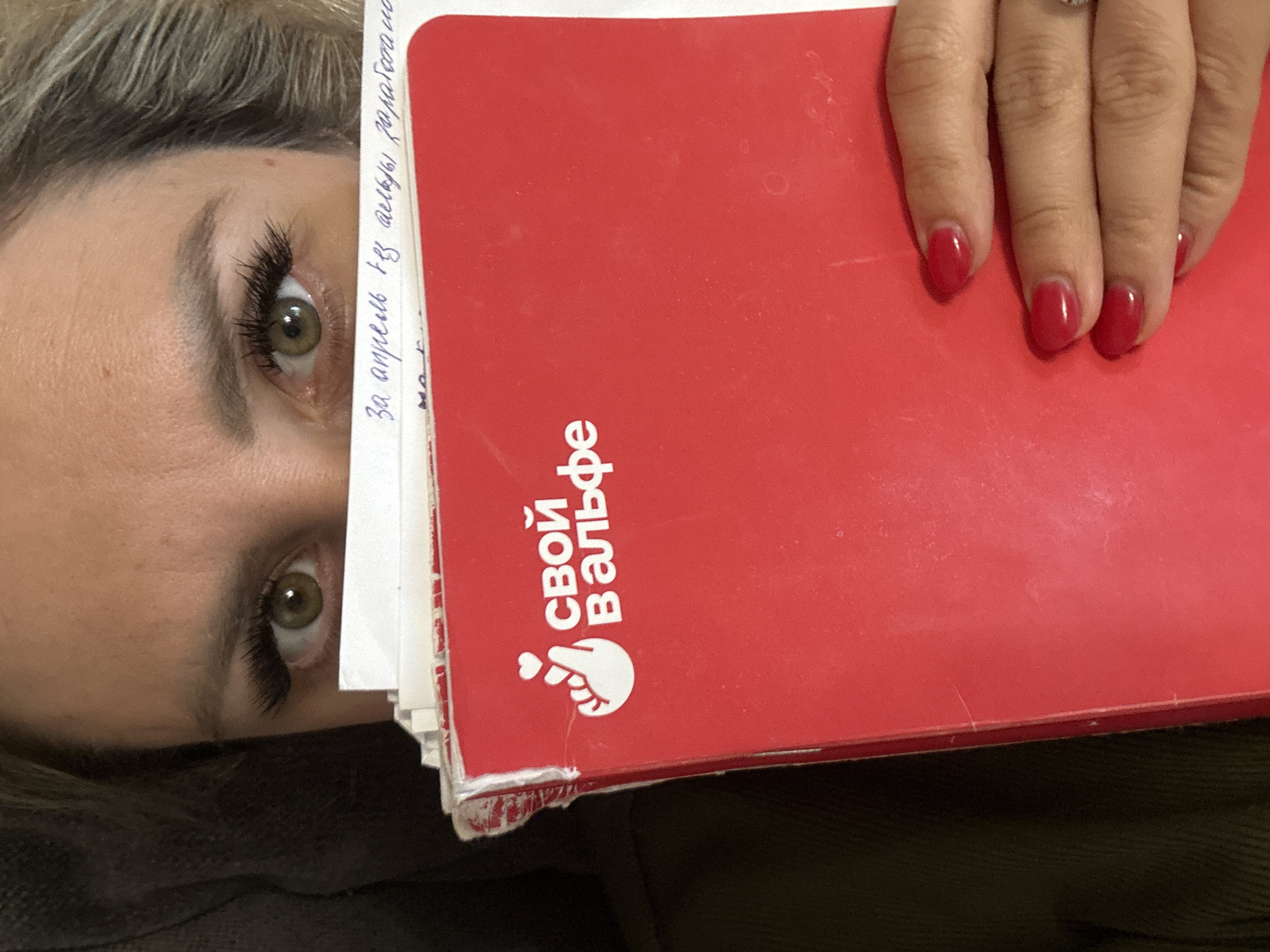 A close-up, overhead shot captures a person's eye peeking out from behind a bright red notebook, with their hand resting on the notebook. The person has light brown hair and striking green eyes with dramatic false eyelashes. The notebook has white pages with handwritten text in blue ink on the left side, partially obscured by the notebook's cover. The text appears to be in Russian, with the phrase "За апрель из Альфы" (For April from Alfa) being partially visible. On the red cover, white text reads "свой ВАЛЬФ" (your WALF) and a small graphic of a hand holding a heart. The person's fingernails are painted a vibrant red. The scene is likely indoors, given the lighting, and the context suggests a personal or professional setting where the notebook is being used for notes or planning. The overall mood is intimate and curious, with the partial reveal of the face drawing the viewer's attention to the eye. The red notebook and matching nail polish add a bold and eye-catching element to the composition.