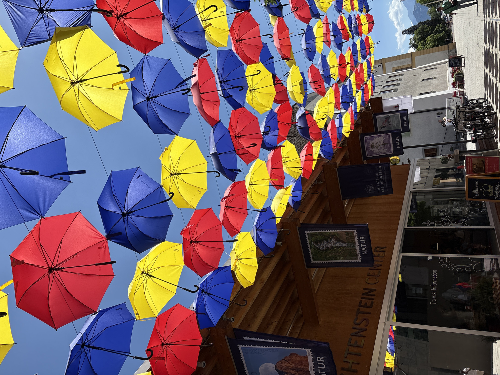 This media file captures a street scene in Vaduz, Liechtenstein, adorned with a vibrant canopy of colorful umbrellas suspended overhead. Red, blue, and yellow umbrellas are strung across the sky, creating a festive and eye-catching display against a clear blue sky, suggesting a bright, sunny day.
Below the umbrellas, the architecture of Vaduz is visible, with buildings featuring wooden facades and glass windows. Signs with text like "LICHTENSTEIN CENTER," "NATUR," and "Tourist Information" indicate commercial or informational establishments. The umbrellas are artfully arranged, some partially obscuring the buildings and sidewalks.
The image also shows glimpses of people, though they are not the primary focus. A few individuals are seen walking on the paved street, and bicycles are parked near the buildings. The overall atmosphere appears to be that of a pleasant day in a European town, with the umbrella installation adding a cheerful and artistic element to the urban landscape. The presence of postage stamp-themed banners, such as "PHILATELIE LIECHTENSTEIN," further reinforces the location in Liechtenstein.
