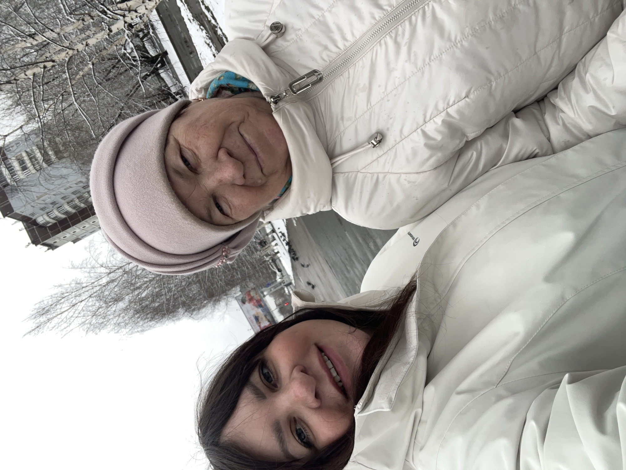 This rotated image captures a selfie of two women, an older woman and a younger woman, outdoors in a snowy, urban environment, likely in Perm, Russia.
The older woman, positioned in the upper part of the frame, has a warm, gentle smile and is looking towards the camera. She wears a light-colored, possibly pale pink or beige, knitted hat and a light cream or white quilted winter jacket. A patterned blue and white fabric lining is visible around her neck. The zipper pull on her jacket features a silver accent and a visible brand logo that reads "OVENTY" with a circular graphic.
Below her, the younger woman, with dark hair, also smiles at the camera. She wears a smooth, light cream or white winter jacket, which also displays the "OVENTY" logo with a circular graphic on its upper left chest area.
The background reveals bare trees sparsely covered in snow, indicating a winter season. Through the branches, apartment buildings and what appears to be a street or pathway can be seen. The overall lighting is soft, suggesting an overcast daytime. Both women appear cheerful and comfortable, dressed warmly for the cold weather.