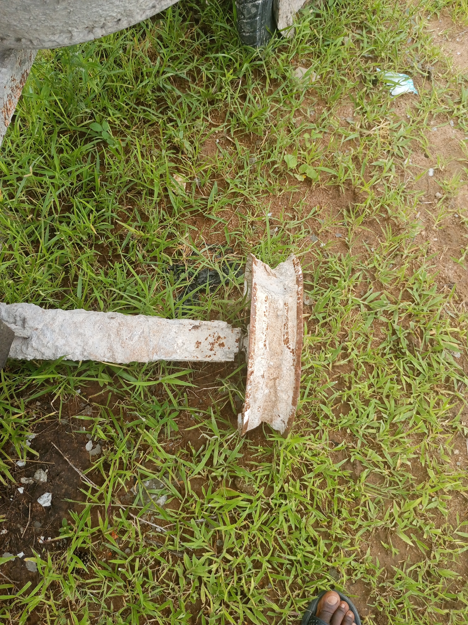 The image shows a close-up, high-angle view of a section of ground covered in green grass and brown soil. In the foreground, the toe and foot of a person wearing a dark sandal are visible. A white, weathered, and rusted metal object lies horizontally across the grassy area, with a piece of grey, rough material on the left and a U-shaped section of rusted metal on the right. The U-shaped metal piece is broken and appears to be part of a larger structure. The overall scene suggests an outdoor environment, possibly a yard or undeveloped area, in Jalingo, Nigeria. The lighting indicates it is daytime, and the presence of grass suggests a season with sufficient rainfall. There is no clear activity or event taking place, as the image is a still shot of objects and a person's foot. No visible text is present in the image.