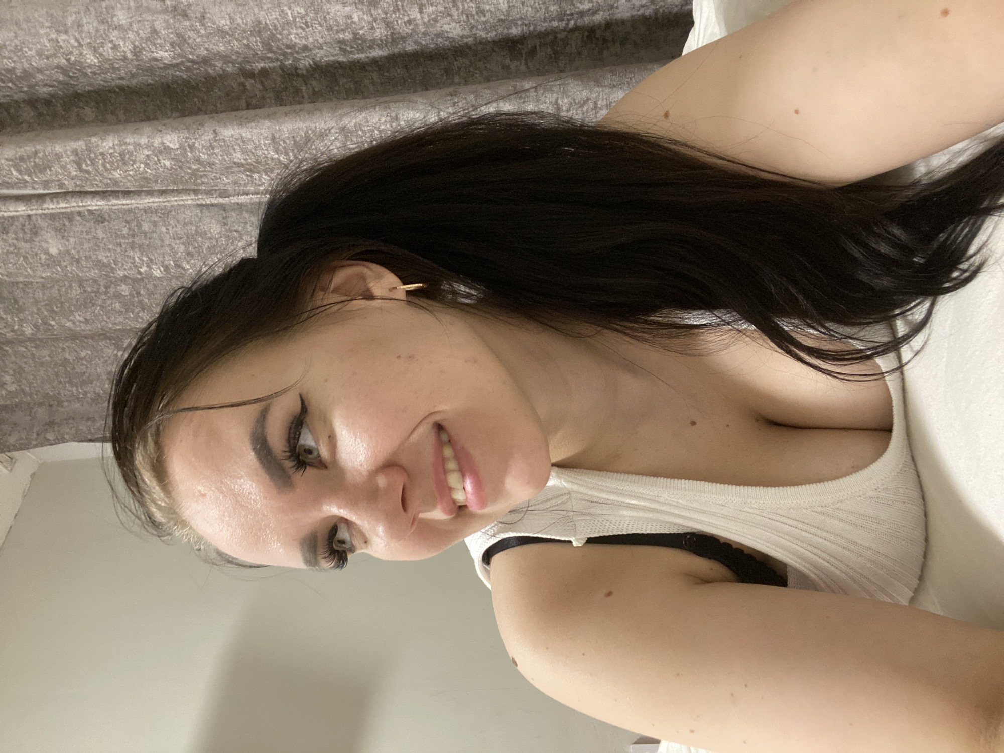 A woman with long dark hair pulled back into a ponytail is lying on a white surface. She is wearing a white tank top with black straps showing. The background consists of a neutral-colored wall and draped fabric. The location is not discernible in the image. The city of Chaikovsky, Russia, cannot be confirmed as the location based on the provided image.
