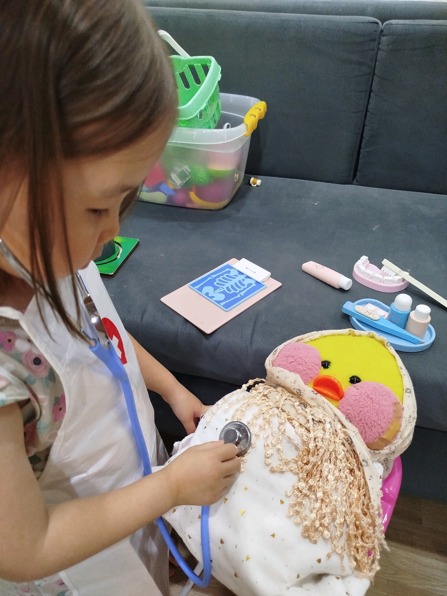A young girl, seen from the side and in partial profile, is engaged in imaginative play. She is wearing a white apron over a floral-print dress and has a blue stethoscope around her neck, with one earpiece in her ear and the diaphragm of the stethoscope placed on a large, plush stuffed duck. The duck is yellow with pink cheeks, black eyes, and an orange beak, and is wrapped in a light-colored fabric with gold speckles and a decorative fringe. The scene takes place indoors, likely in a living room, with the girl and the duck positioned on a dark grey sofa.
Scattered on the sofa around the duck are various toy medical accessories. To the right of the duck, there is a pink toy dental mold with teeth and a toothbrush. Next to that, a blue tray holds a toy thermometer displaying "39°C," and two small bottles. Further left, there's a pink tube that resembles a toy toothpaste or ointment, and a light pink rectangular object with a blue medical graphic on it, possibly a toy X-ray. In the background, a clear plastic bin filled with colorful toys sits next to a bright green plastic basket. The girl's left arm is visible, and her right hand is holding the stethoscope diaphragm. Her hair is brown and pulled back. The lighting suggests an indoor environment, possibly with natural light from a window or overhead lighting. The setting does not provide any specific visual cues to indicate the city of Mirny, Russia.