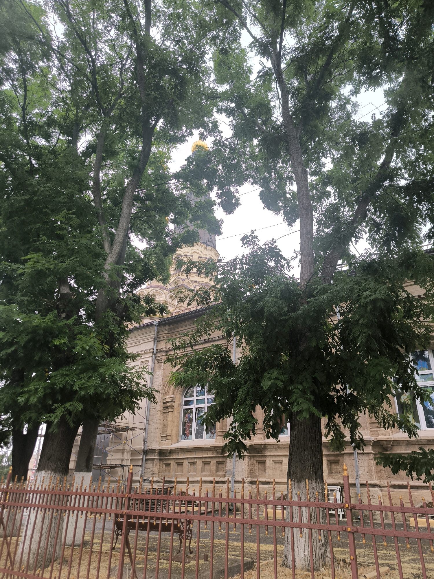 This image depicts the exterior of the Church of the Assumption of the Blessed Virgin Mary, located at Soborna Street 18, Mykolaiv, Ukraine. The church, built in the Neo-Byzantine style, features a prominent golden dome visible above the trees. The building's facade is constructed from light-colored stone, with arched windows and decorative elements. Trees with lush green foliage frame the view of the church, and a red metal fence with golden finials runs along the foreground. Scaffolding is visible on the left side of the church, suggesting ongoing renovation or construction work. The sky is overcast, casting a soft, diffused light on the scene. No people are visible in the image.