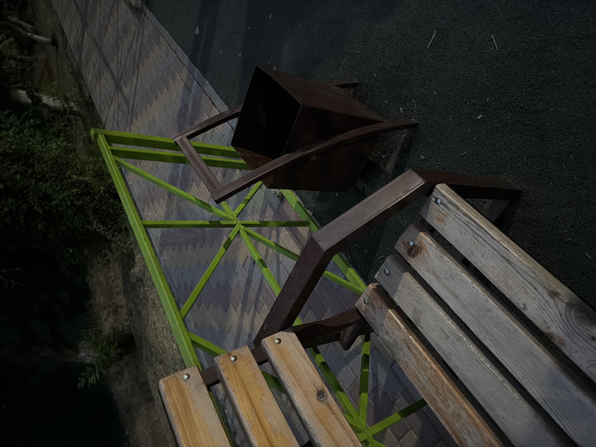 The image depicts a park bench and a trash can located in Shymkent, Kazakhstan. The scene is set outdoors, likely in a public park, as indicated by the presence of a paved walkway, greenery, and a fence. The bench is made of weathered wood and has a metal frame, appearing to be slightly aged. Adjacent to the bench is a dark-colored metal trash can with a distinctive angular design. A bright green metal fence runs along the edge of the walkway, separating it from the grassy area. The lighting suggests it is either dusk or a dimly lit area of the park. There are no people visible in the image. The environment appears calm and undisturbed.