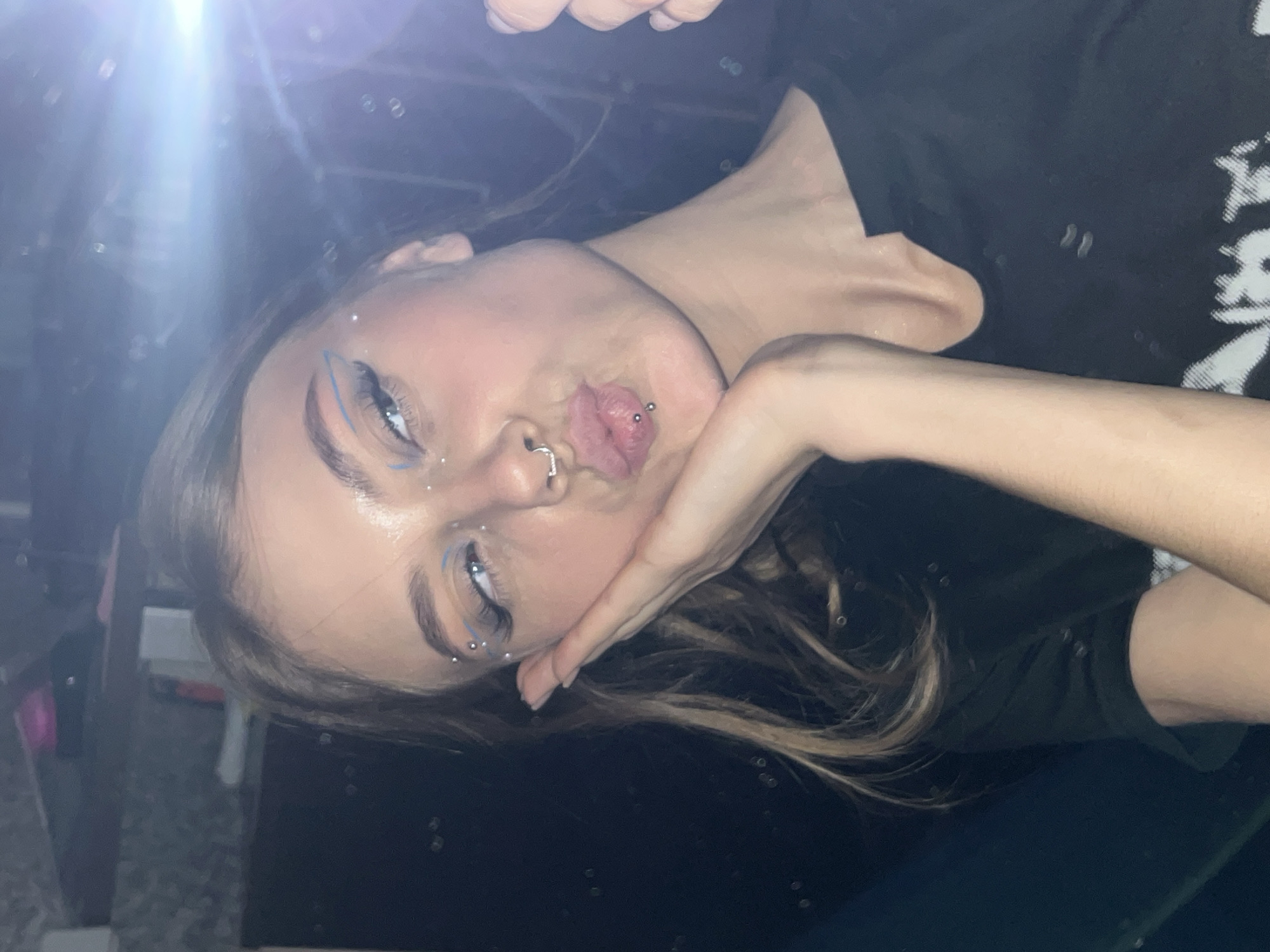 A young woman with a lip piercing and a nose ring is posing for a photo. She has blue eyeliner and glitter on her face. She is wearing a black t-shirt. She is holding her hand up to her face and puckering her lips. The background is dark and out of focus. The photograph was not taken in Фоки, Russia.