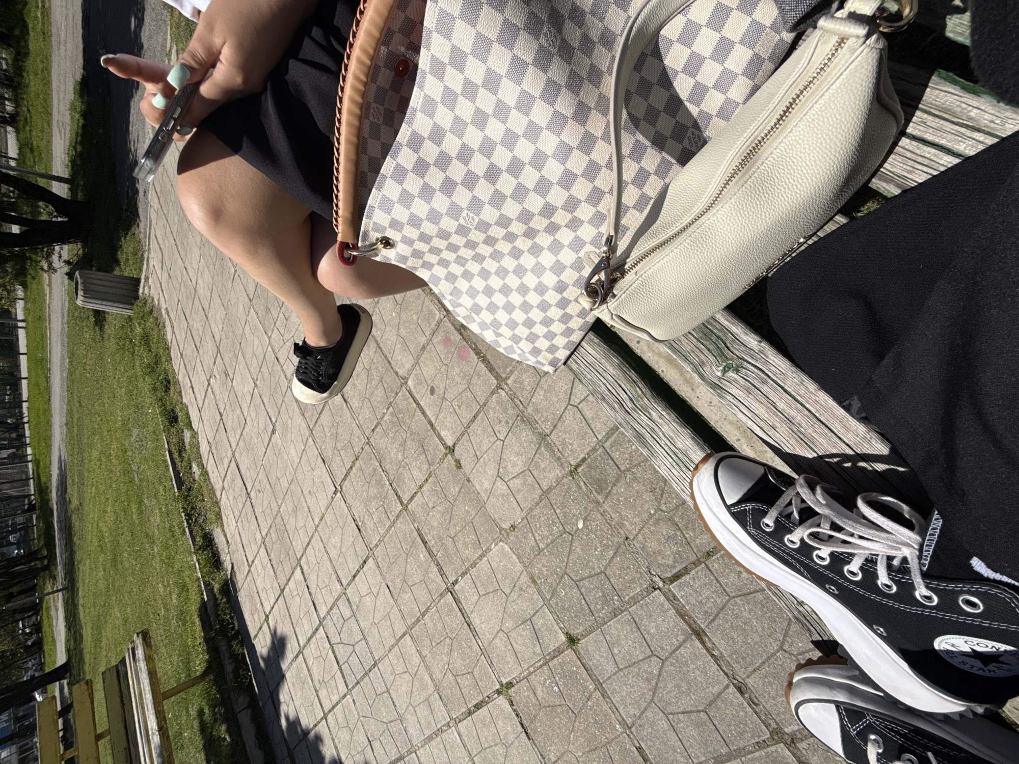 The image captures a ground-level perspective of two individuals seated outdoors, likely on a bench in a public space. One person is holding a smartphone and wears black shoes with a white sole, while the other individual wears black Converse sneakers. A large checkered pattern bag with tan leather trim and a smaller white bag sit on the bench between them. The ground is covered in paving stones with patches of grass visible on the side. Given the style of the paving and the overall setting, it's possible this photo was taken in Sofia, Bulgaria.