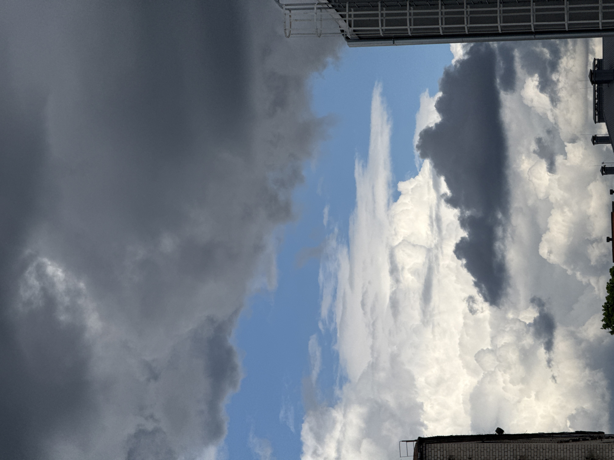The image shows a sky view from Riga, Latvia. A distinct contrast of dark, ominous storm clouds on the left half dominates the scene, juxtaposed against a brighter, sunlit section with fluffy white clouds and patches of blue sky on the right. Man-made structures, possibly rooftops and building elements, are visible at the top and bottom of the frame, providing a sense of urban context. The presence of ventilation or similar fixtures is noted on one of the buildings. Greenery appears at the right corner, suggesting vegetation amidst the urban landscape.